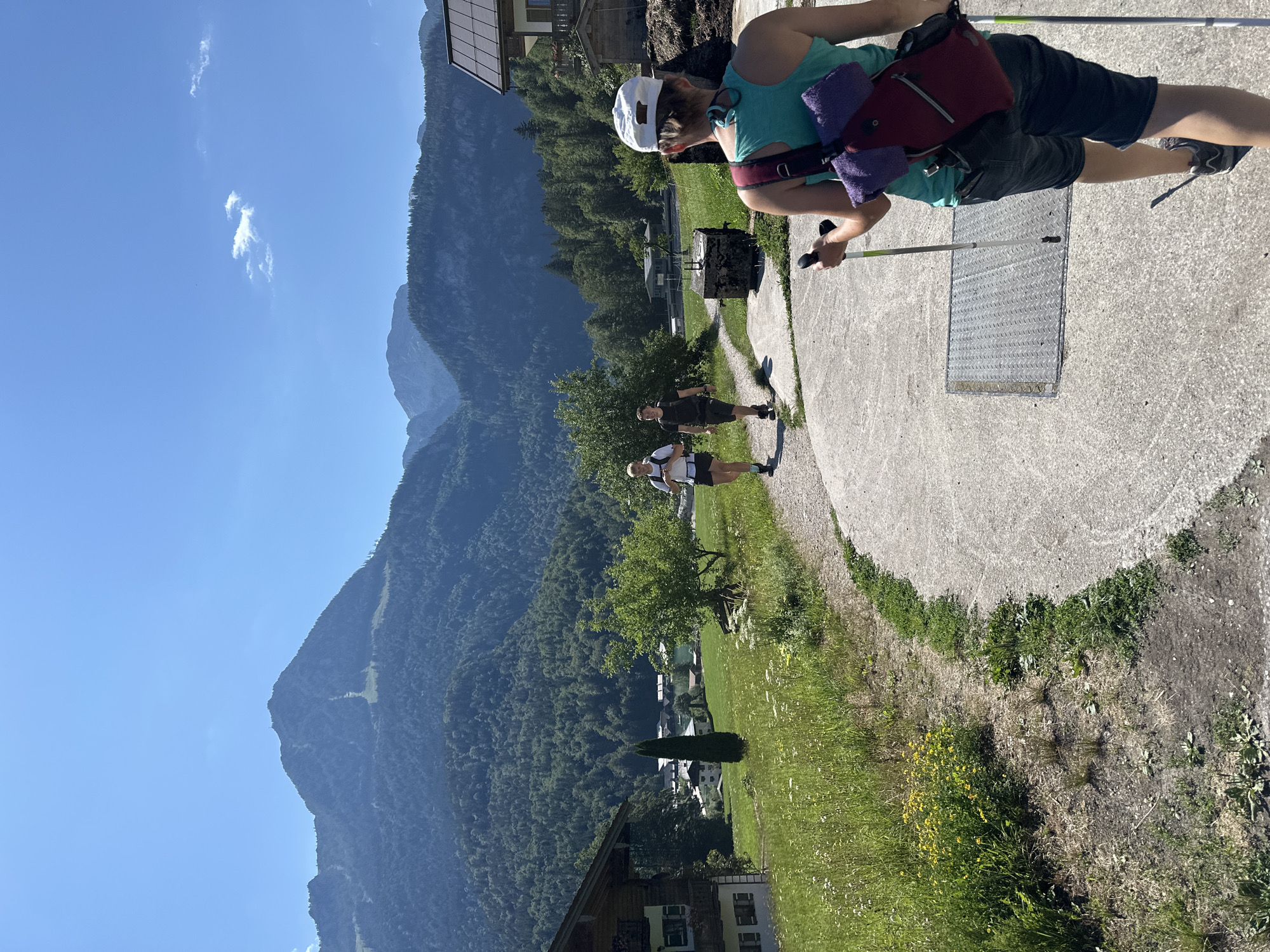 In Schönau am Königssee, Germany, on a clear, sunny day, two individuals are walking away from the camera on a paved path. The person in the foreground, wearing a white baseball cap, turquoise tank top, and dark shorts, is using trekking poles. A red backpack with a purple towel draped over it is slung over their shoulder. Behind them, two other individuals, both appearing to be male, are walking on the same path. They are wearing athletic clothing and backpacks. The path winds through a grassy area with wildflowers and trees. In the background, a lush green mountain covered in dense forest rises towards a bright blue sky. Several traditional Bavarian-style houses with wooden facades are visible nestled among the trees and meadows. The scene depicts a scenic hiking or walking trail in a mountainous region.