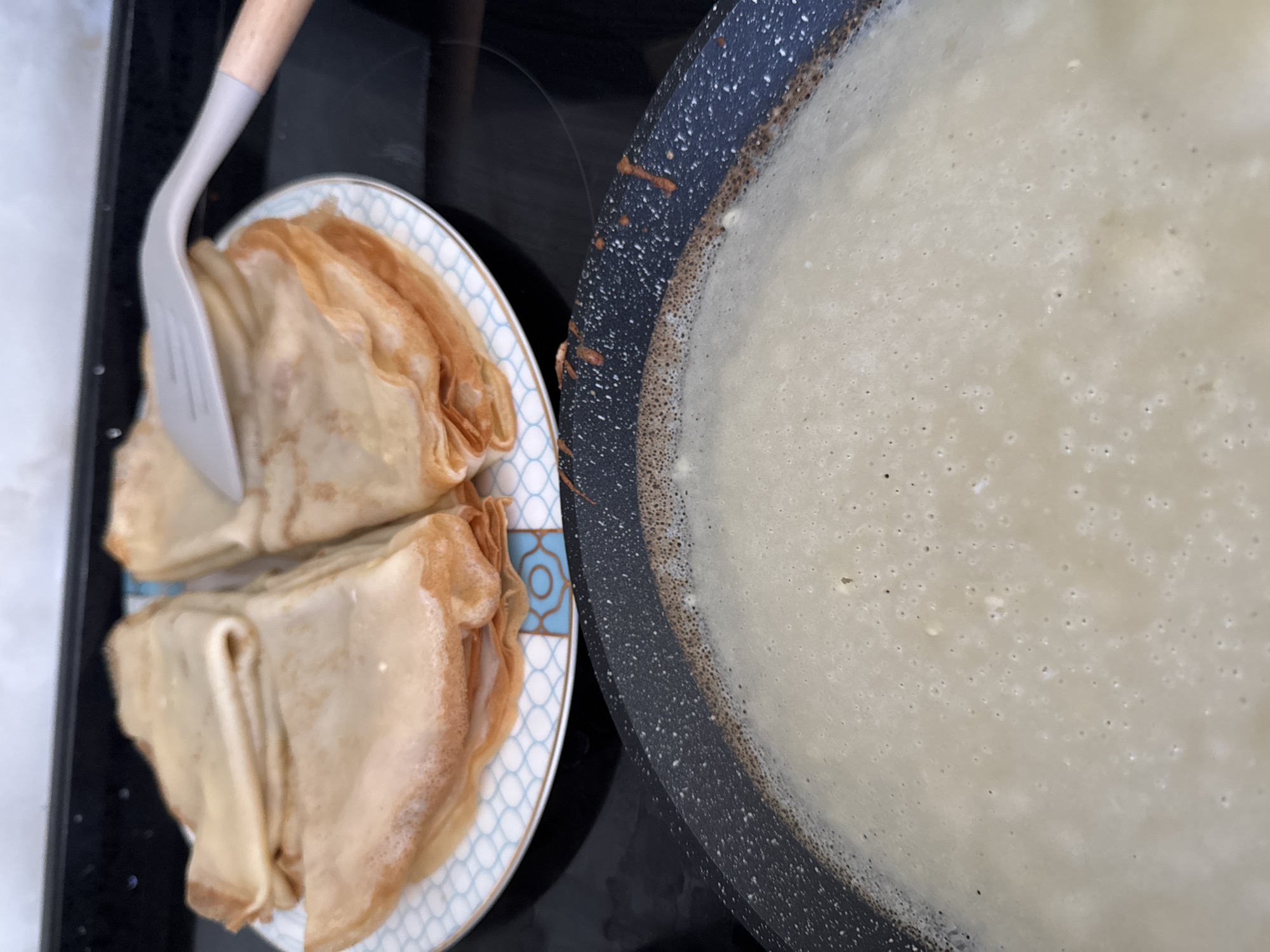 In Uyutnoye, Ukraine, on a kitchen stovetop, a pile of golden-brown crepes is stacked on a white and blue patterned plate. A grey silicone spatula with a wooden handle is partially covering the crepes on the right side. In the foreground, a black non-stick frying pan is filled with a light-colored batter, likely for more crepes, with small bubbles forming on the surface. The background shows a dark stovetop surface.