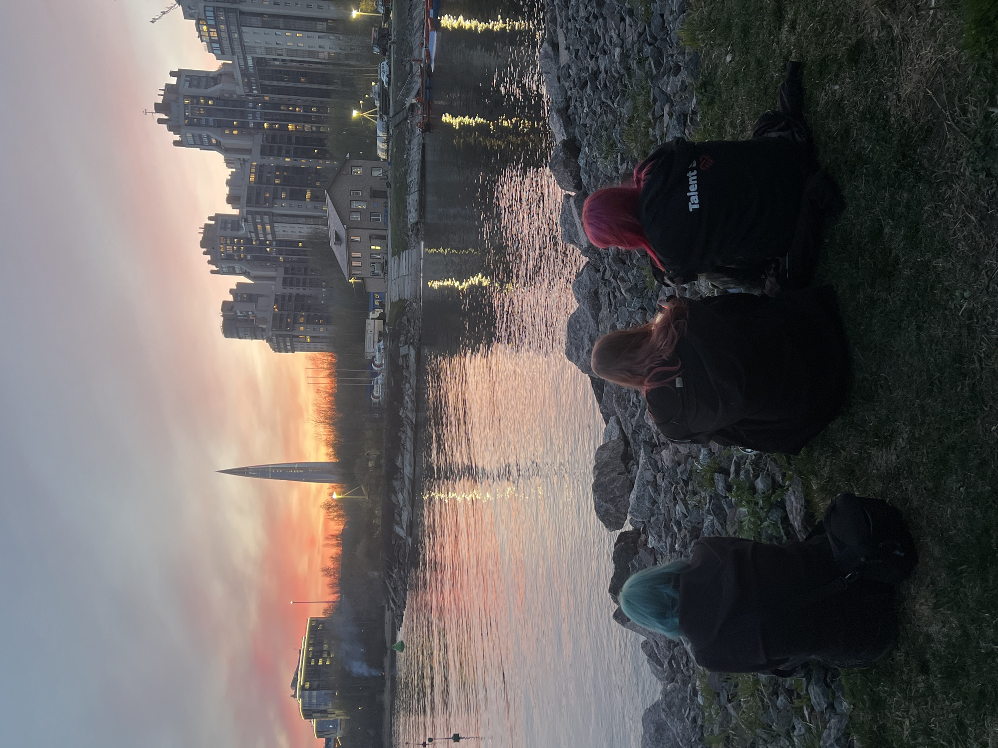 The image depicts a serene sunset scene in Saint Petersburg, Russia, observed from a grassy and rocky embankment. In the foreground, three individuals are seen from behind, sitting or crouching by the edge of a body of water, likely a river or canal. The person on the left has teal-colored hair, the middle person has reddish-brown hair, and the person on the right has bright pink hair. All are wearing dark jackets or hoodies. On the back of the jacket worn by the person with pink hair, the word "Talent" is visible in white letters, alongside a red and white logo or initials that resemble "SJM" or "SJD". One person appears to be holding a phone or camera.
Across the water, a city skyline stretches, featuring numerous residential and commercial buildings, many with illuminated windows, suggesting dusk or early evening. The water reflects the vibrant colors of the sunset sky—pinks, oranges, and purples—as well as the scattered lights from the city. A prominent, tall, slender skyscraper, identifiable as the Lakhta Center, stands out against the colorful sky in the mid-ground. The overall atmosphere is peaceful and contemplative, with the soft, fading light of the day casting a warm glow over the scene.