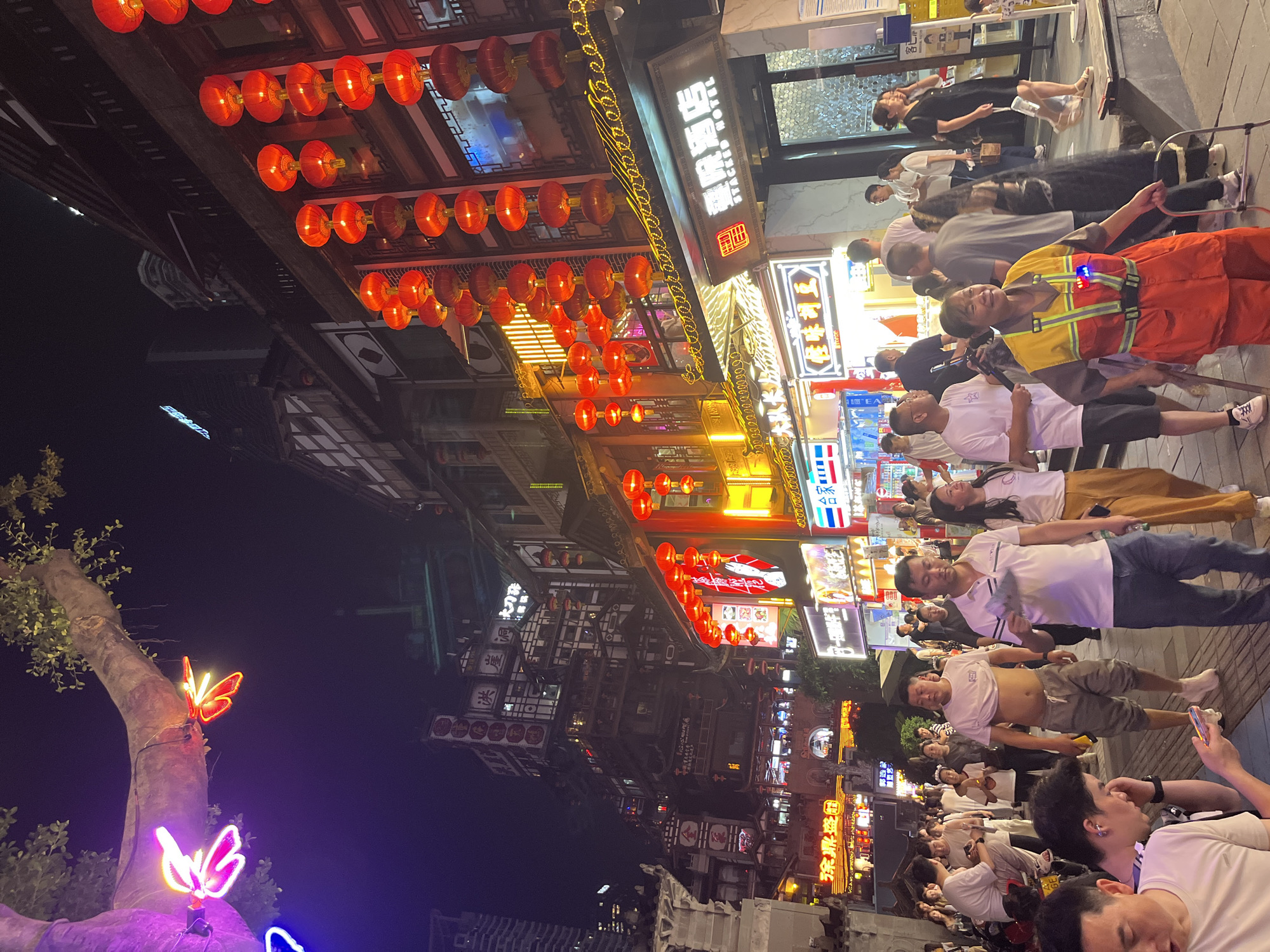 The image depicts a bustling night scene in the Yuzhong District of Chongqing, China. The architecture is a mix of traditional and modern, with buildings adorned with red lanterns and neon signs in Chinese characters. A large tree branch draped with neon lights shaped like butterflies is visible in the upper left corner.
Crowds of people are walking along a street lined with shops and restaurants. Many individuals are dressed casually in t-shirts and pants. Some are looking at their phones, while others are engaged in conversation. A woman in a bright orange vest and a woman in a black dress are particularly prominent in the foreground. The ground is paved with stone tiles. The overall atmosphere is lively and vibrant, characteristic of a popular urban tourist destination at night.