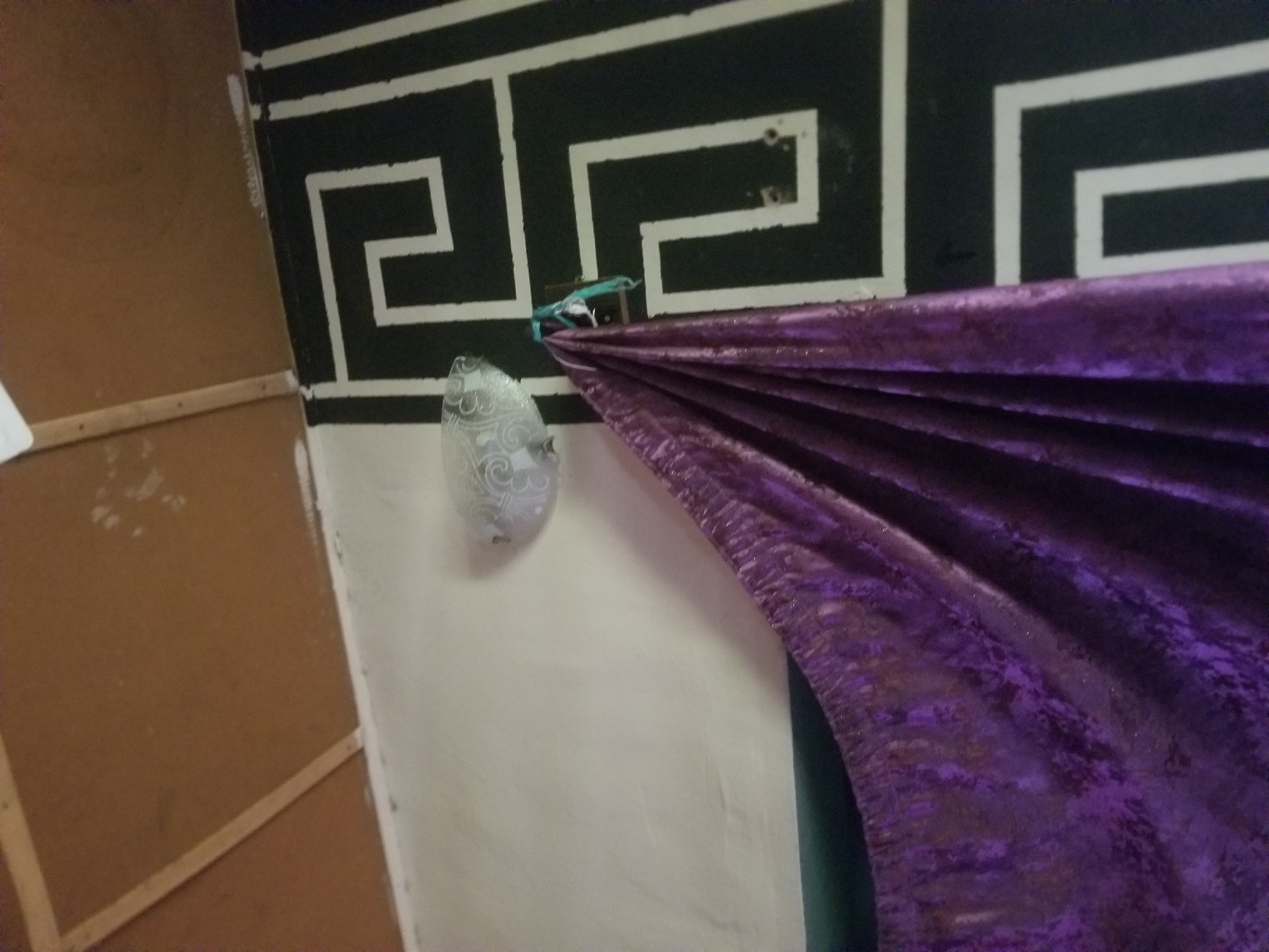 The image shows a close-up view of a wall decorated with a black and white geometric pattern. A piece of purple, shimmering fabric is draped over the wall, obscuring part of the pattern. The fabric is gathered and pleated, creating a textured appearance. To the left of the fabric, a small, oval-shaped object with a swirl pattern is attached to the wall. This object appears to be made of a translucent material, possibly glass or plastic. A teal-colored cord or string is used to attach the purple fabric to the wall, near the oval object. The wall itself is predominantly white, with some visible imperfections or marks. To the left of the white wall, a section of a brown, wooden surface is visible, possibly a panel or partition, with some horizontal wooden strips attached to it. The lighting suggests an indoor setting, possibly artificial light, as there are no strong shadows indicating natural sunlight. The overall scene appears to be a decorative arrangement or a backdrop, possibly for an event or performance. The image is static, with no discernible activity taking place beyond the way the fabric is draped. The location context is provided as Jauro Yinu, Nigeria, but this information cannot be directly inferred from the visual elements in the image.