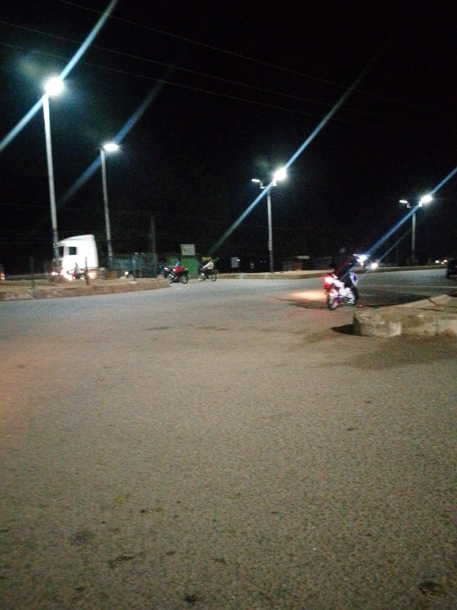 This image depicts a nighttime scene at a road intersection or roundabout in Bukuru, Nigeria. The environment is illuminated by multiple overhead streetlights with bright, possibly LED, fixtures, casting strong light and noticeable lens flares across the dark sky. The road surface is asphalt, appearing worn and textured.
In the mid-ground, a white semi-trailer truck cab is visible on the left side of the road, facing towards the right. Several individuals are operating motorcycles on the road. Most prominently, a motorcycle with an illuminated red taillight is positioned in the mid-ground right, angled towards the left, with a single rider. Further into the intersection, other motorcycles with riders are observed, one moving from right to left, and two others appearing smaller and further back on the left side of the road. Concrete curbs or low barriers delineate parts of the intersection. Overhead power lines are faintly visible against the dark sky. No direct interaction between individuals is discernible.
