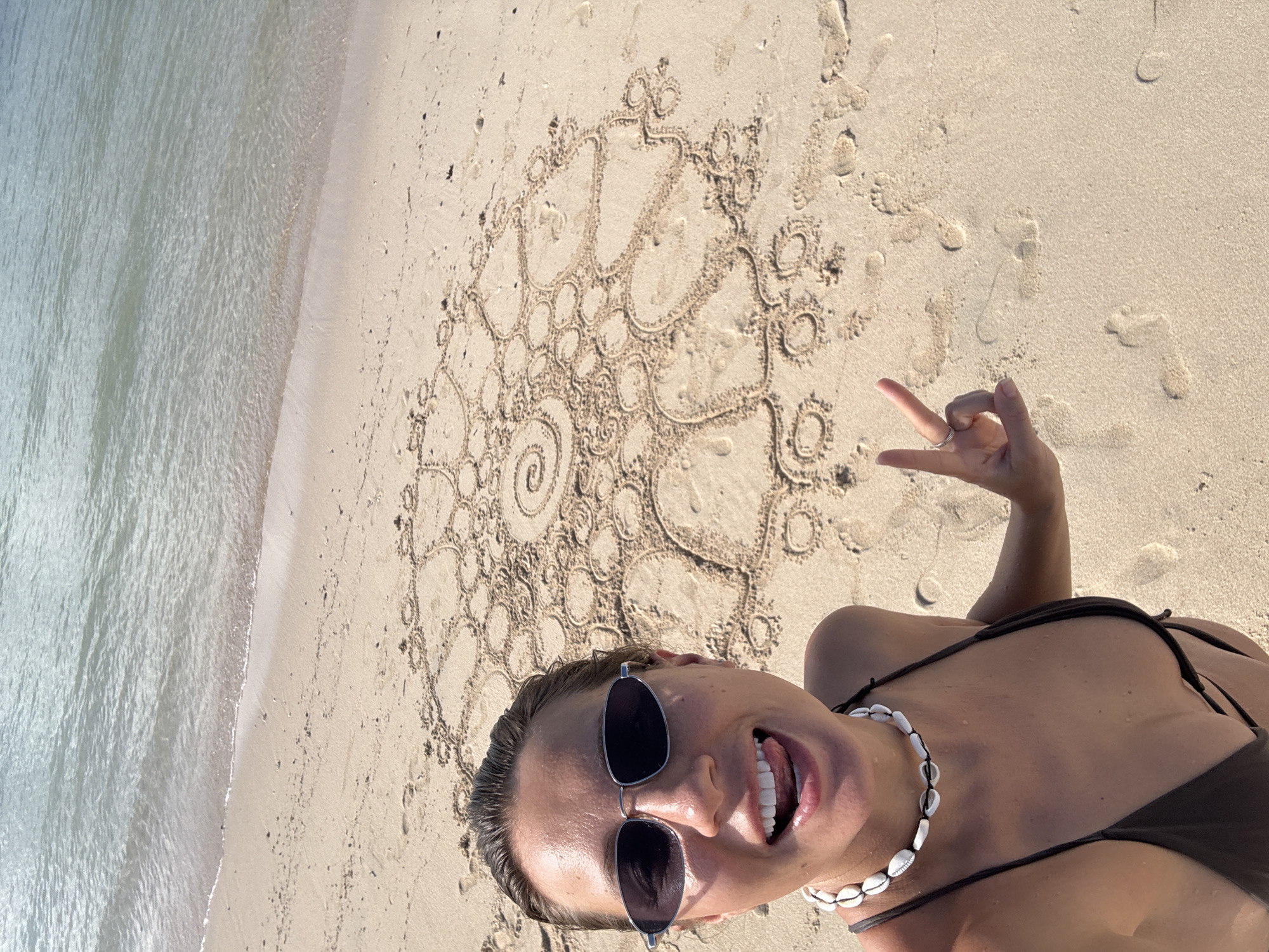 A woman is on a beach in Kuta Selatan, Indonesia. She is wearing sunglasses and a shell necklace. She is wearing a brown bikini. She is smiling and making a hand gesture. A complex pattern is drawn in the sand behind her. The ocean is visible in the background. The beach is sandy.