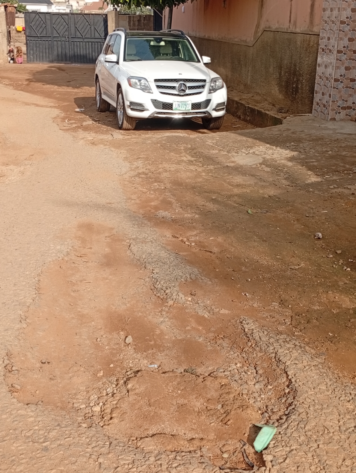 In Rafin Par, Nigeria, a white Mercedes-Benz GLK-Class SUV is parked on a dirt road. The vehicle, with license plate "IJN 775CF", faces the viewer from a slightly elevated angle. The road surface is rough and uneven, composed of compacted dirt and loose stones, with visible erosion and potholes. In the background, a large black metal gate stands open, revealing a glimpse of a building behind it. To the left of the gate, two figures, possibly children, are partially visible. To the right of the vehicle, a textured wall with a patterned tile section suggests the presence of a building. The overall environment appears to be an outdoor urban setting characterized by unpaved roads and modest structures.