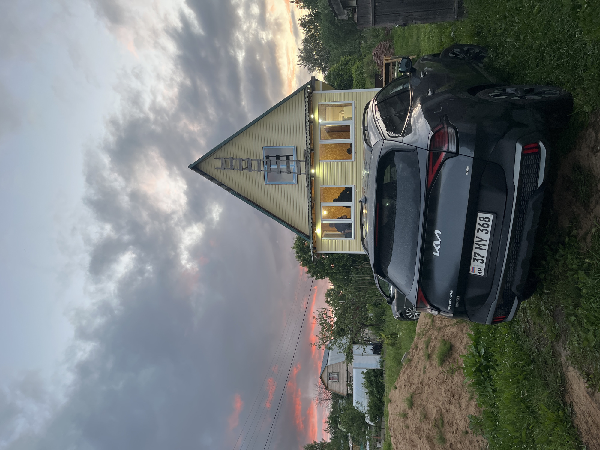In Клобушнево, Russia, a dark gray Kia Sportage is parked on a grassy area in front of a two-story yellow house. The house has several windows illuminated from within and a ladder leaning against its peaked roof. Behind the house, more trees and a greenhouse are visible. A large pile of sand is to the left of the house, and another car is parked in the background. The sky is dramatic with dark clouds streaked with vibrant pink and orange hues from a sunset or sunrise. The scene appears to be a rural residential area.