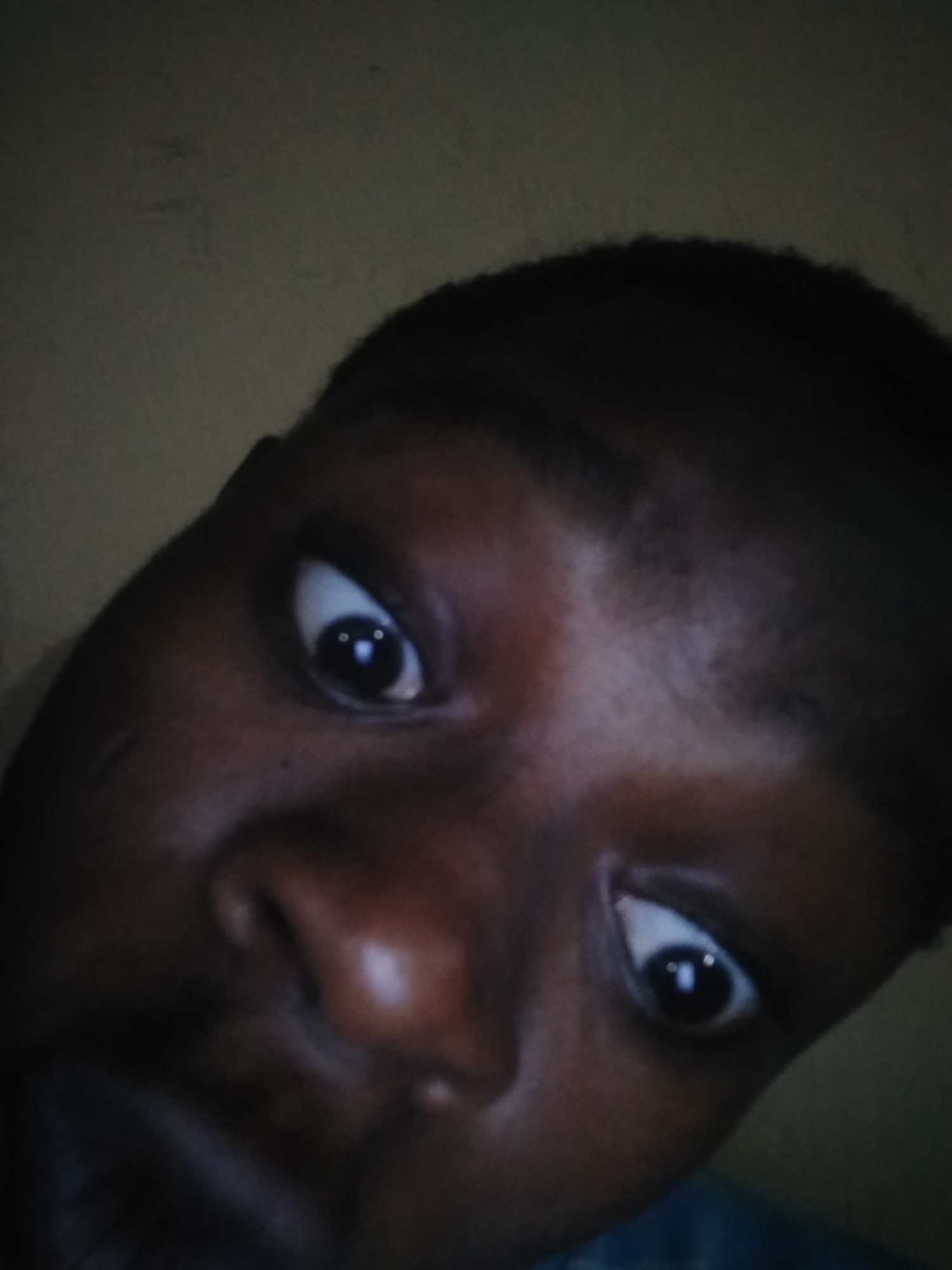 A close-up, low-angle selfie of a dark-skinned individual with short, dark hair. The person's eyes are wide and directed towards the camera. The background appears to be a dimly lit interior space with a plain wall. The provided location of Shangisha, Nigeria cannot be verified from the image alone.