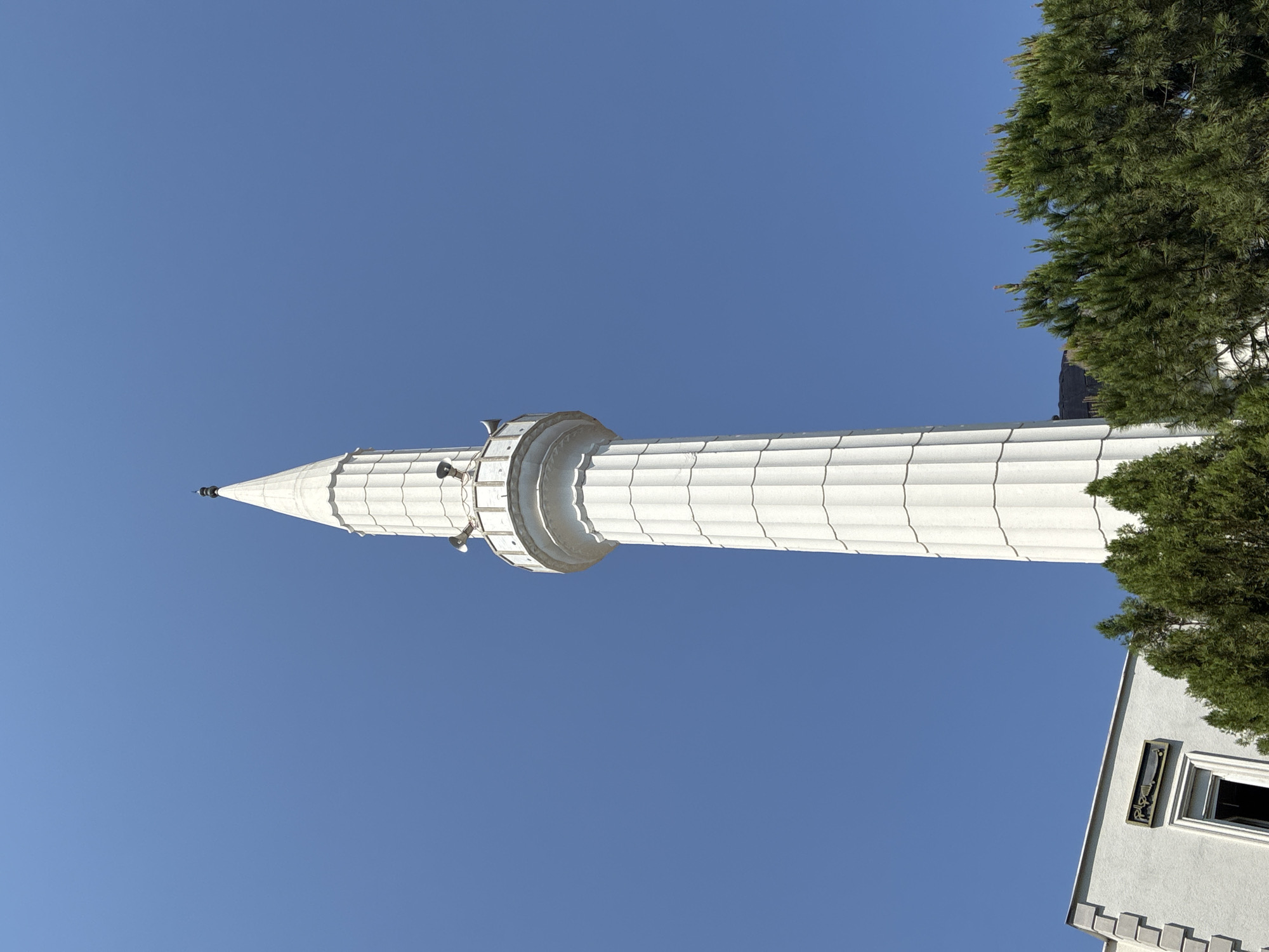 The image shows the minaret of a mosque in Alanya, Turkey, against a clear blue sky. The minaret is tall and white, with a fluted design and a pointed top. It has a balcony near the top, with loudspeakers attached. In the foreground, there are green trees and part of a white building with an inscription. The building appears to be attached to the mosque. The image is taken from a low angle, looking up at the minaret.