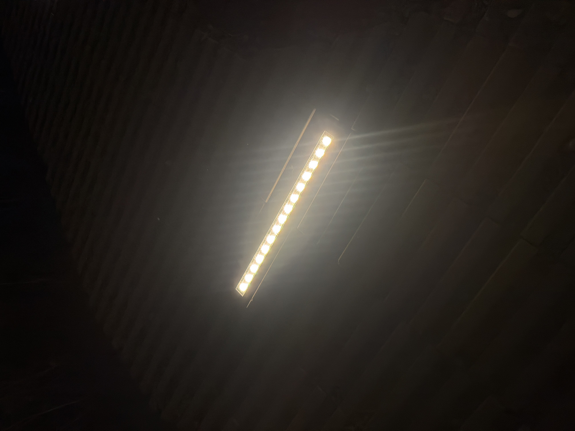 An illuminated LED light bar, composed of multiple individual bulbs, is positioned diagonally across a dark, textured surface. The light bar emits a bright, warm white light that causes lens flare and a diffuse glow in the surrounding darkness. The surface below appears to be made of dark, elongated elements, possibly wood planks or tiles, laid in a parallel pattern. The overall scene is dimly lit, suggesting an outdoor environment at night or in a poorly lit indoor space. There are no people or discernible actions visible in the image. The location in Shymkent, Kazakhstan, is not discernible from the visual elements alone.