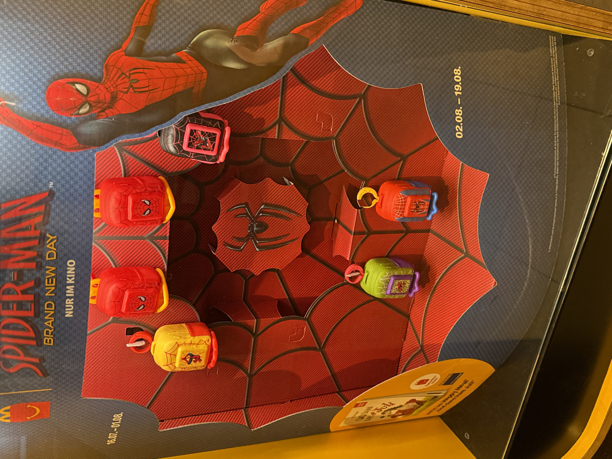 The image depicts a promotional display for a Spider-Man themed McDonald's Happy Meal, likely located in Saalfeld/Saale, Germany. The display features a large cardboard cutout of Spider-Man in his iconic red and blue suit, dynamically posed. In the foreground, arranged on a red, spiderweb-patterned backdrop, are several small, collectible toy backpacks. These toys, designed as mini backpacks, each feature a Spider-Man motif. Some are predominantly red, while others are yellow or green, with varying details and accessories. Text on the display includes "SPIDER-MAN", "BRAND NEW DAY", "NUR IM KINO" (Only in cinemas), and dates "16.07.-01.08." and "02.08. - 19.08.". A portion of a yellow McDonald's promotional stand is visible on the left, with text that appears to be in German, including "Happy Meal" and potentially information about the promotion. The overall presentation suggests a point-of-sale display within a McDonald's restaurant.