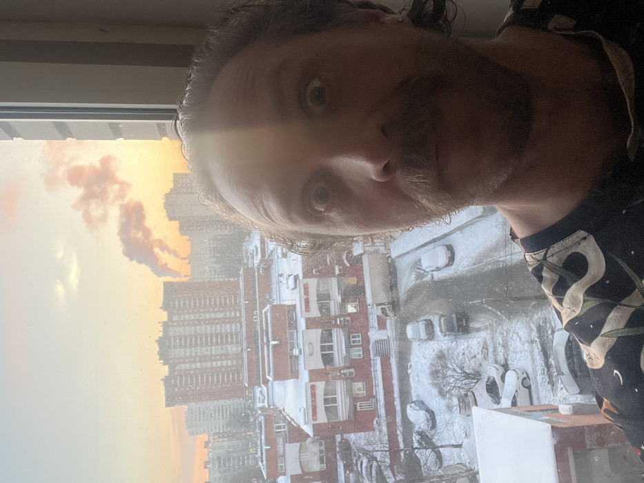 An adult male with light complexion, curly brown hair, and a prominent mustache is captured in a selfie from an indoor window view. His eyes are wide, and he exhibits a surprised or exaggerated expression. A strong, vertical band of direct sunlight illuminates the right side of his face and forehead, casting the left side into relative shadow. He wears a dark t-shirt adorned with a light-colored, patterned design.
The scene outside the window, viewed from an elevated position, depicts a snow-covered urban landscape in Saint Petersburg, Russia, under what appears to be a sunrise or sunset, characterized by warm orange and pink sky hues. Light snowfall is visible throughout the frame. Immediately below, a two-story red brick commercial building with its roof covered in snow features several windows, and a partially visible Cyrillic sign reads "СТРОИТЕЛЬНЫЕ" (Construction). A snow-covered road or parking area winds through the foreground, populated by several parked cars, also covered in snow, and a white delivery truck. Barren trees and shrubs are scattered on the snowy ground. In the mid-ground and background, numerous multi-story residential apartment buildings, indicative of post-Soviet architecture, dominate the skyline. Further in the distance, prominent industrial chimneys emit large plumes of reddish-pink smoke, illuminated by the low sun, suggesting an industrial zone within this residential area of Saint Petersburg.