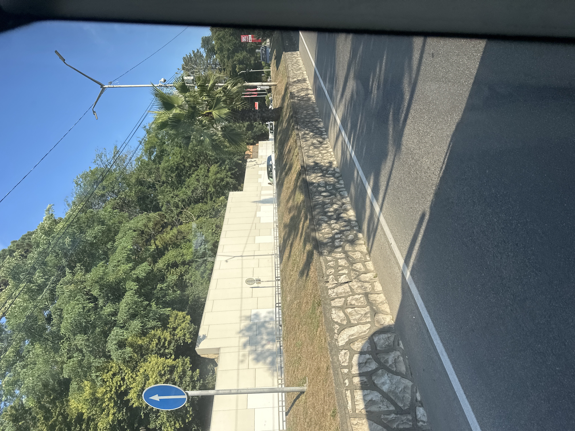 The image is a photograph taken from a moving vehicle on a sunny day in Sochi, Russia. The road is asphalt with a white line marking the lane. To the left of the road, there is a retaining wall made of natural stone, topped with a grassy embankment. Behind the embankment is a long, pale concrete wall. A blue traffic sign with a white arrow pointing upwards indicates that this is a one-way street. Trees and foliage grow on a hillside behind the concrete wall. A street light and power lines are visible against the clear blue sky. In the distance, there are some buildings and cars, and a palm tree stands near the road. The shadow of the vehicle and the surrounding trees are cast across the road and the retaining wall.