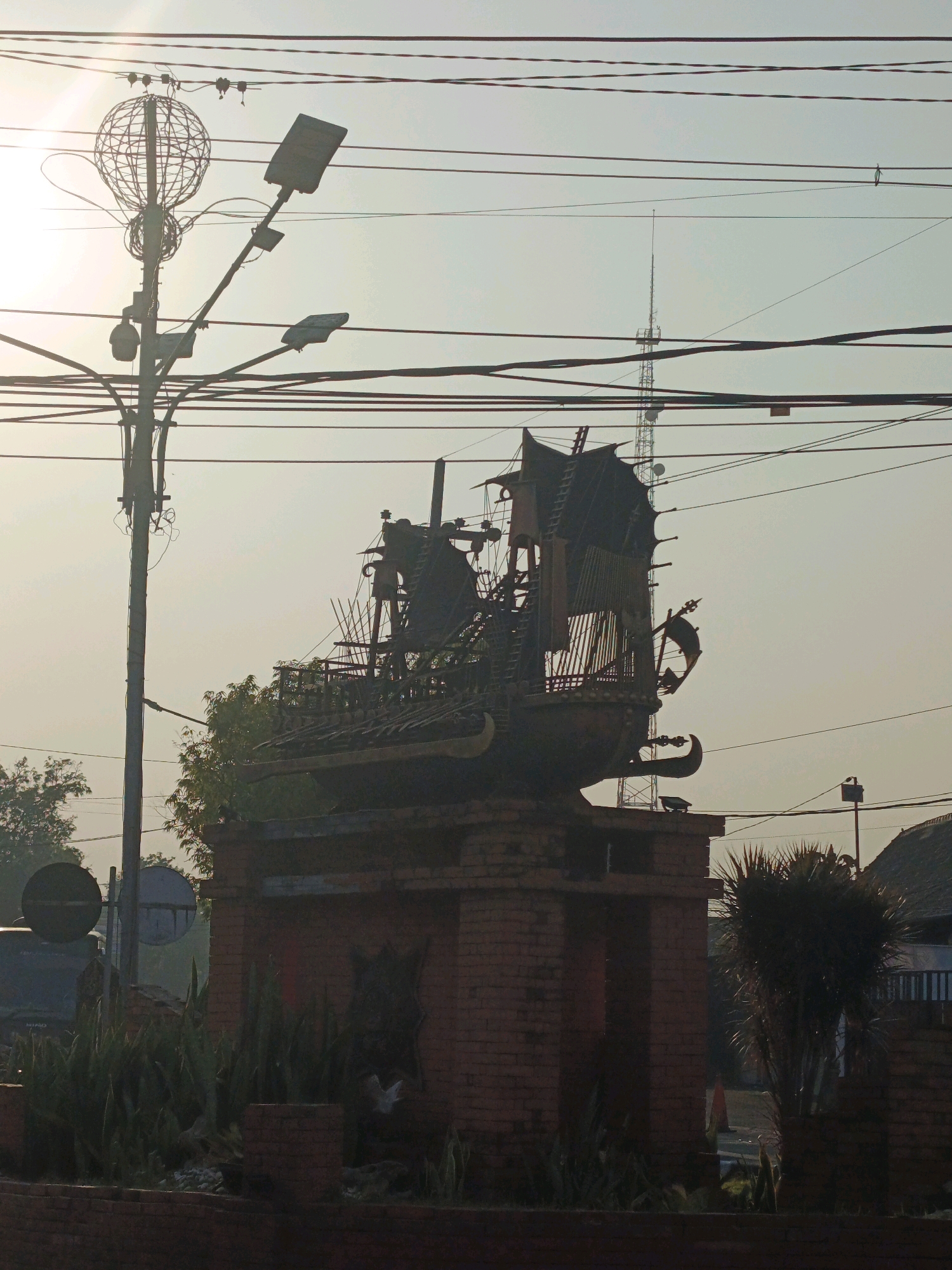 In the city of Miji, Indonesia, a monument resembling a ship is erected on a brick pedestal, its intricate structure crafted from dark metal. The monument is positioned in a public area with several overhead utility lines, including power lines and what appears to be a telecommunication tower in the distance. The scene is illuminated by the soft glow of the sun, which is partially obscured by the utility lines, creating a somewhat hazy atmosphere. Lush green foliage, including tall, spiky plants and trees, surrounds the base of the monument and the surrounding brickwork, adding a touch of nature to the urban landscape. The monument and its surroundings appear to be a landmark or a point of interest within the city.
