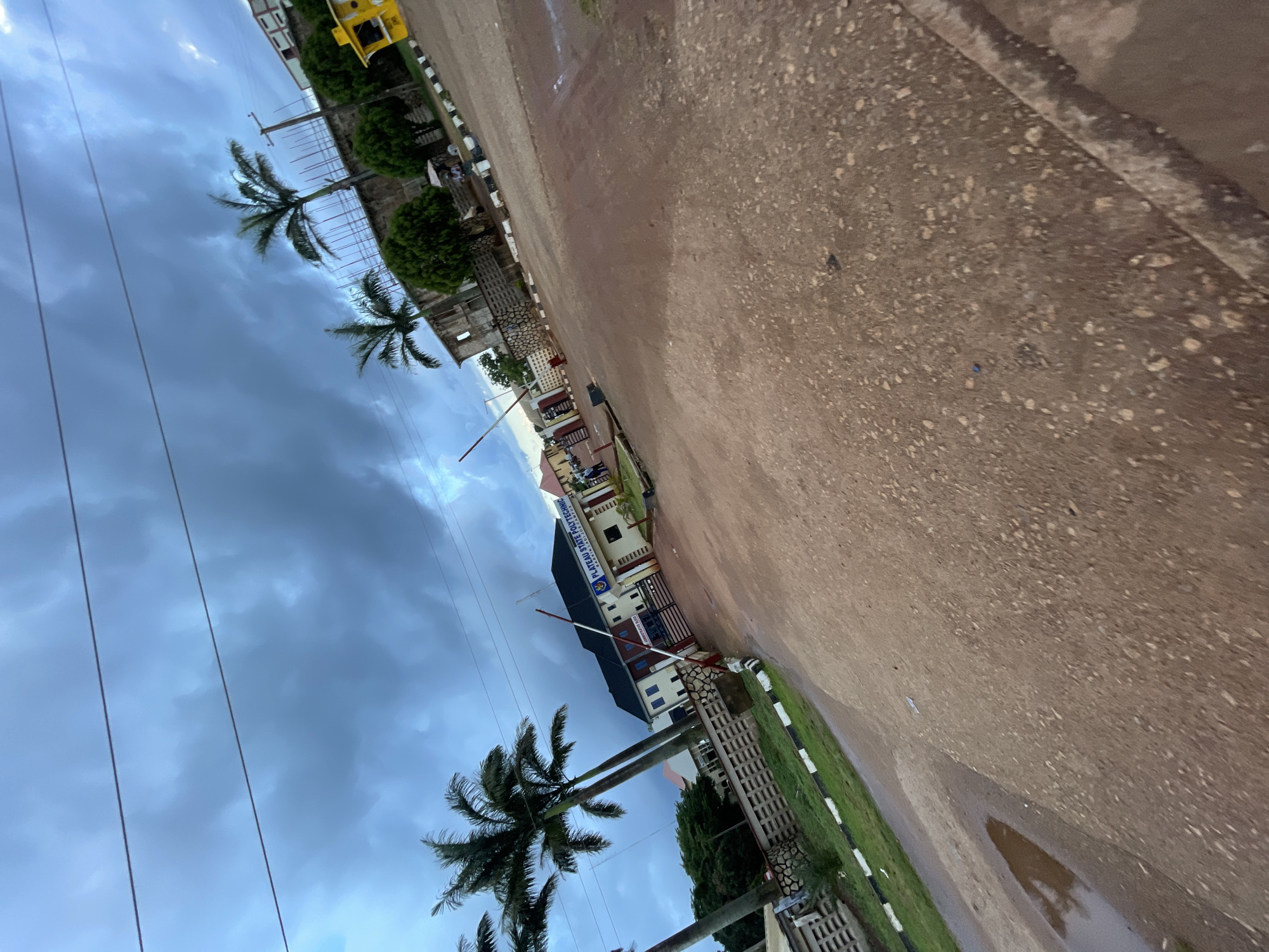 The image shows the entrance to Plateau State Polytechnic in Jos, Nigeria, on a cloudy day. The foreground is dominated by a reddish-brown dirt road, with some puddles of water visible, suggesting recent rain. A white and red barrier arm is partially blocking the road leading into the polytechnic.
The main building visible has "PLATEAU STATE POLYTECHNIC" and "DAMBBA CAMPUS" written on it in blue and white lettering. The building is light-colored with a dark roof and several windows. A black metal gate is situated in front of the building. Palm trees are scattered throughout the scene, with their fronds reaching towards the overcast sky. In the background, there are other buildings and structures, some appearing to be under construction or in disrepair. A yellow security booth is also visible to the right of the frame. There are a few people in the distance near the entrance of the polytechnic. The overall environment appears to be an urban or semi-urban educational institution.