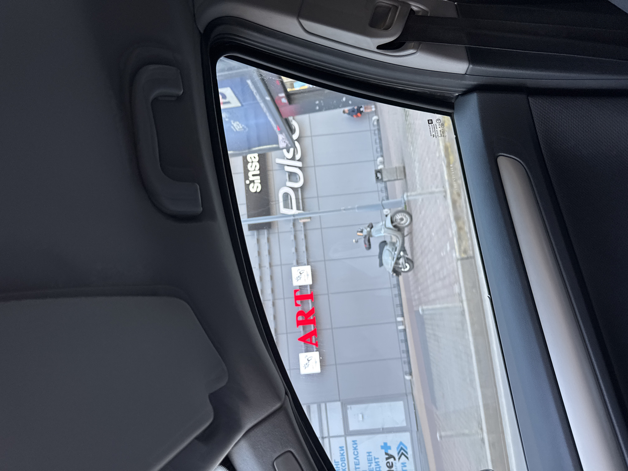 The image shows a view from inside a car, looking out through the side window at a street scene. The location is likely in Sofia, Bulgaria, based on the Cyrillic alphabet used in the signs. A building with the logos "sinsa Pulse" and "ART" is visible, suggesting a commercial or retail space. A scooter is parked on the sidewalk in front of the building. A person is walking further down the street. The car interior includes a grab handle, sun visor, window frame, and upholstery.