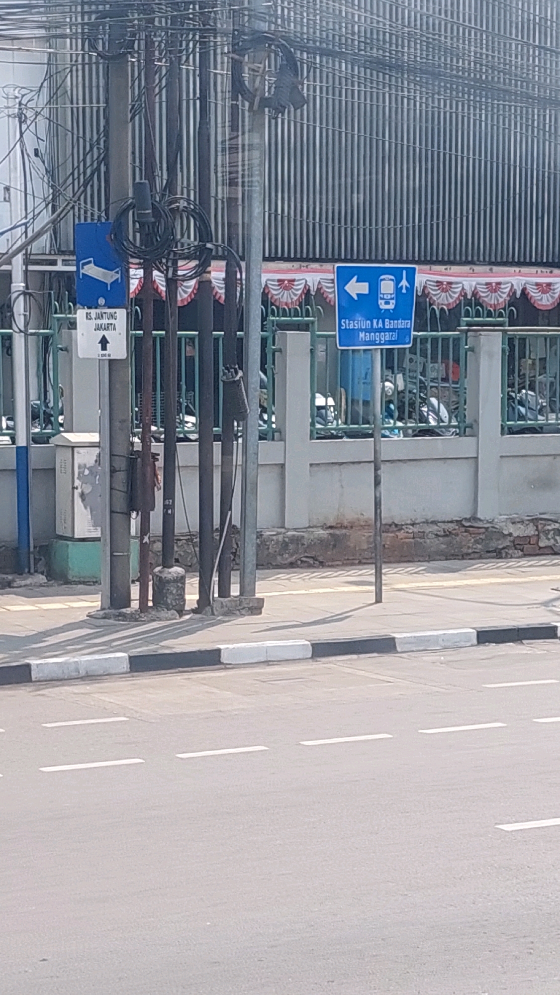 The image depicts a street scene in East Jakarta, Indonesia, featuring directional signage. In the foreground, a gray asphalt road is marked with dashed white lines, indicating lanes of traffic. A concrete sidewalk with a black and white curb runs alongside the road. A utility pole stands on the sidewalk, adorned with numerous electrical wires and cables. Two blue directional signs are attached to the pole. The sign on the left shows a white bed icon and the text "RS. JANTUNG JAKARTA" with an upward arrow, indicating the direction to the Jakarta Heart Hospital. The sign on the right displays a white arrow pointing left, accompanied by icons of a train and an airplane, and the text "Stasiun KA Bandara Manggarai," directing towards the Manggarai Airport Train Station. Behind the sidewalk, a green metal fence encloses an area with a light-colored building and parked motorcycles visible. The building has a facade of vertical gray slats. The overall environment suggests an urban setting with functional infrastructure and traffic-related information.