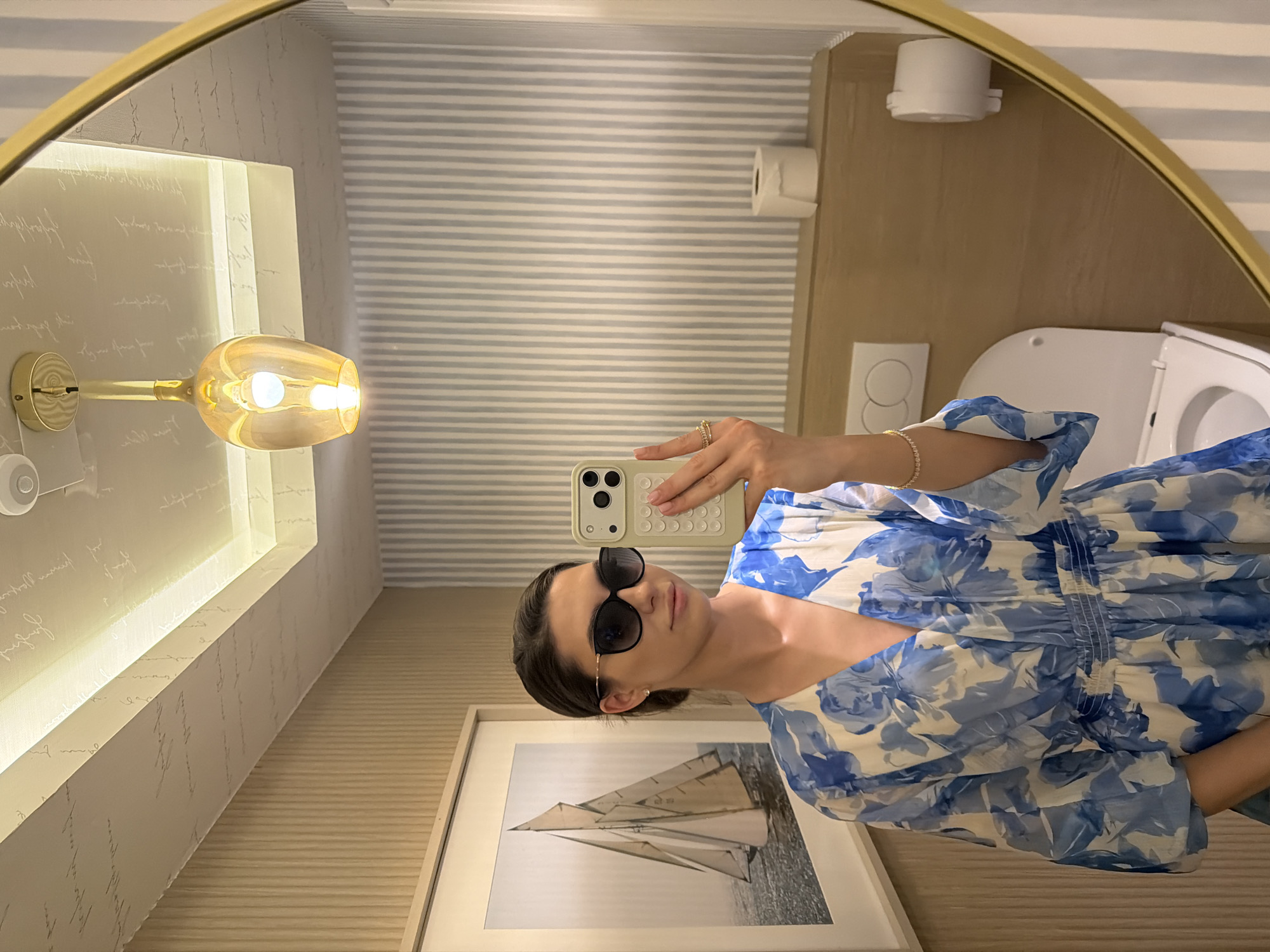 A woman wearing sunglasses and a blue floral dress is taking a selfie in a bathroom. The bathroom has horizontally striped walls, a toilet, a toilet paper dispenser, and a framed picture of a sailboat. There is also a light fixture on the left side of the mirror, and the mirror has a gold frame. The location is likely Rafailovići, Montenegro, based on the metadata of similar images.