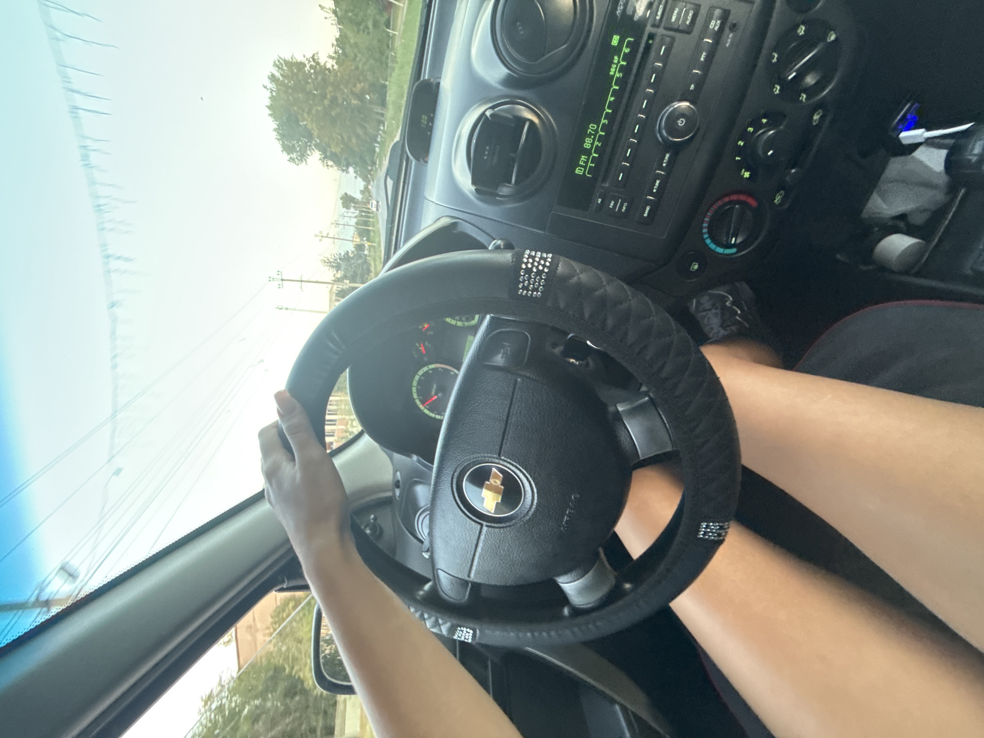 The image shows a person's legs and hands on the steering wheel of a car. The car's interior is visible, including the dashboard, radio, and air conditioning controls. The steering wheel has a black cover with diamond-like accents. The person is driving on a road with trees and buildings visible on either side. The sky is clear and blue. The image appears to have been taken during the daytime. There is no specific information in the image to suggest the location is Abaclia, Moldova.