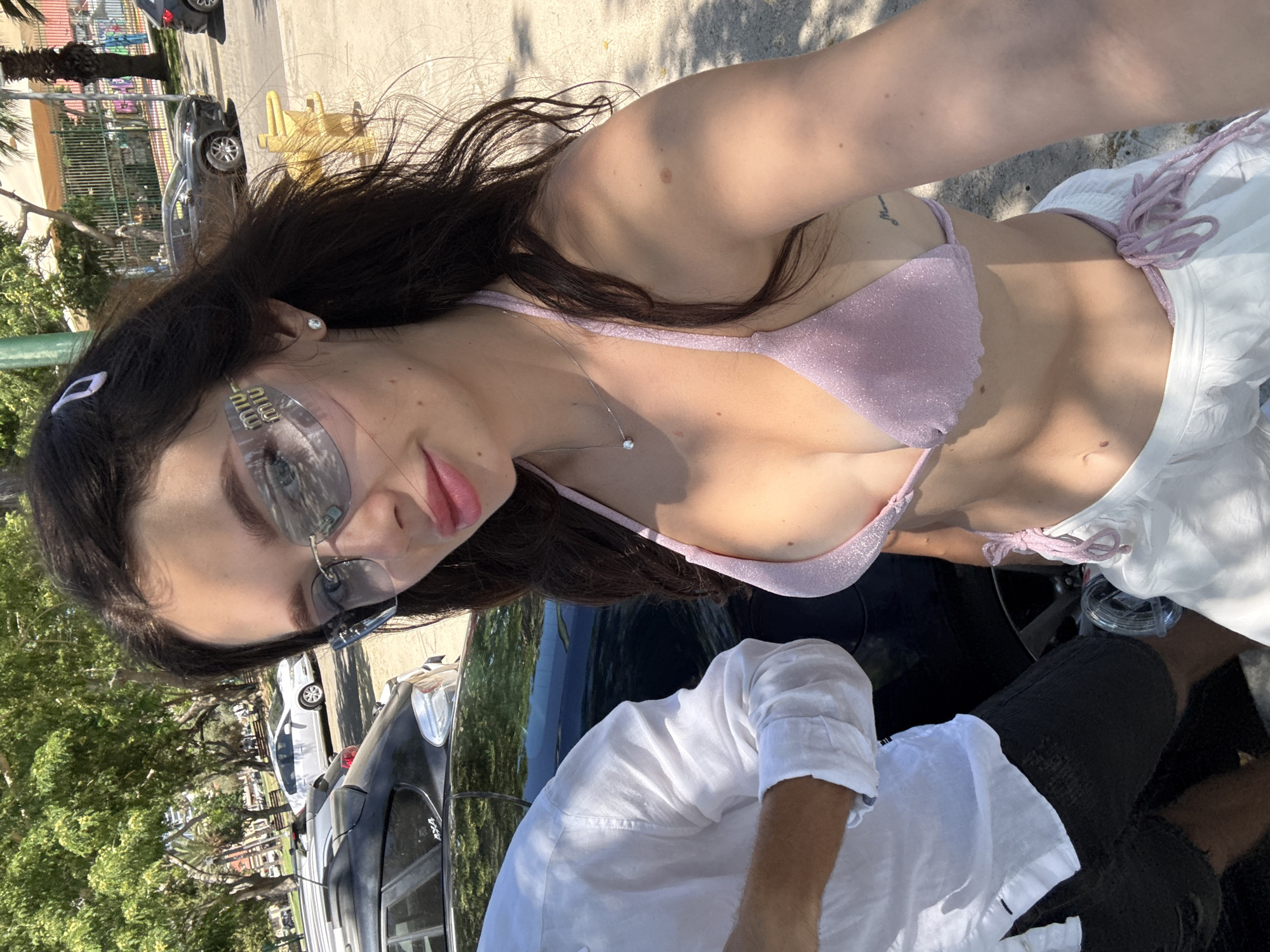 A young woman with dark hair, wearing sunglasses and a pink bikini top, takes a selfie in Tel Aviv, Israel. She is standing next to a man wearing a white shirt and dark shorts. Cars and trees are visible in the background, suggesting an outdoor, urban setting. The overall scene appears casual and summery.