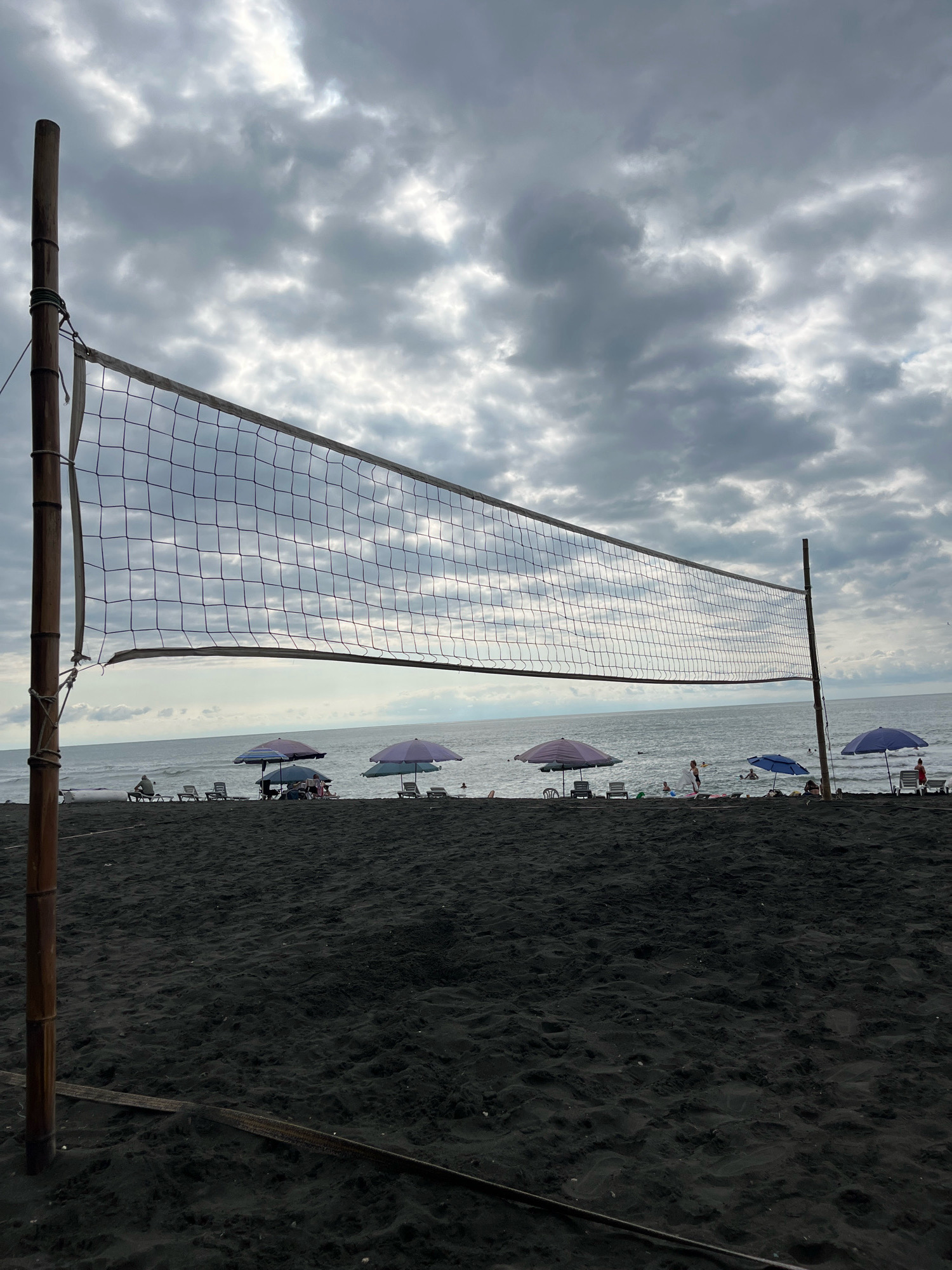 A foreground shot of a black sand beach in Ureki, Georgia, features a volleyball net supported by bamboo poles. The net spans the width of the frame, with the ocean visible beyond it. The sky is overcast with dramatic, cloudy formations. In the mid-ground, several lounge chairs and colorful umbrellas are arranged along the shoreline. A few people are scattered on the beach, some seated in chairs, others standing near the water. The ocean has moderate waves, and a few individuals are visible in the water, suggesting swimming or recreational activity. The overall scene conveys a sense of a cloudy, yet potentially active, beach day.