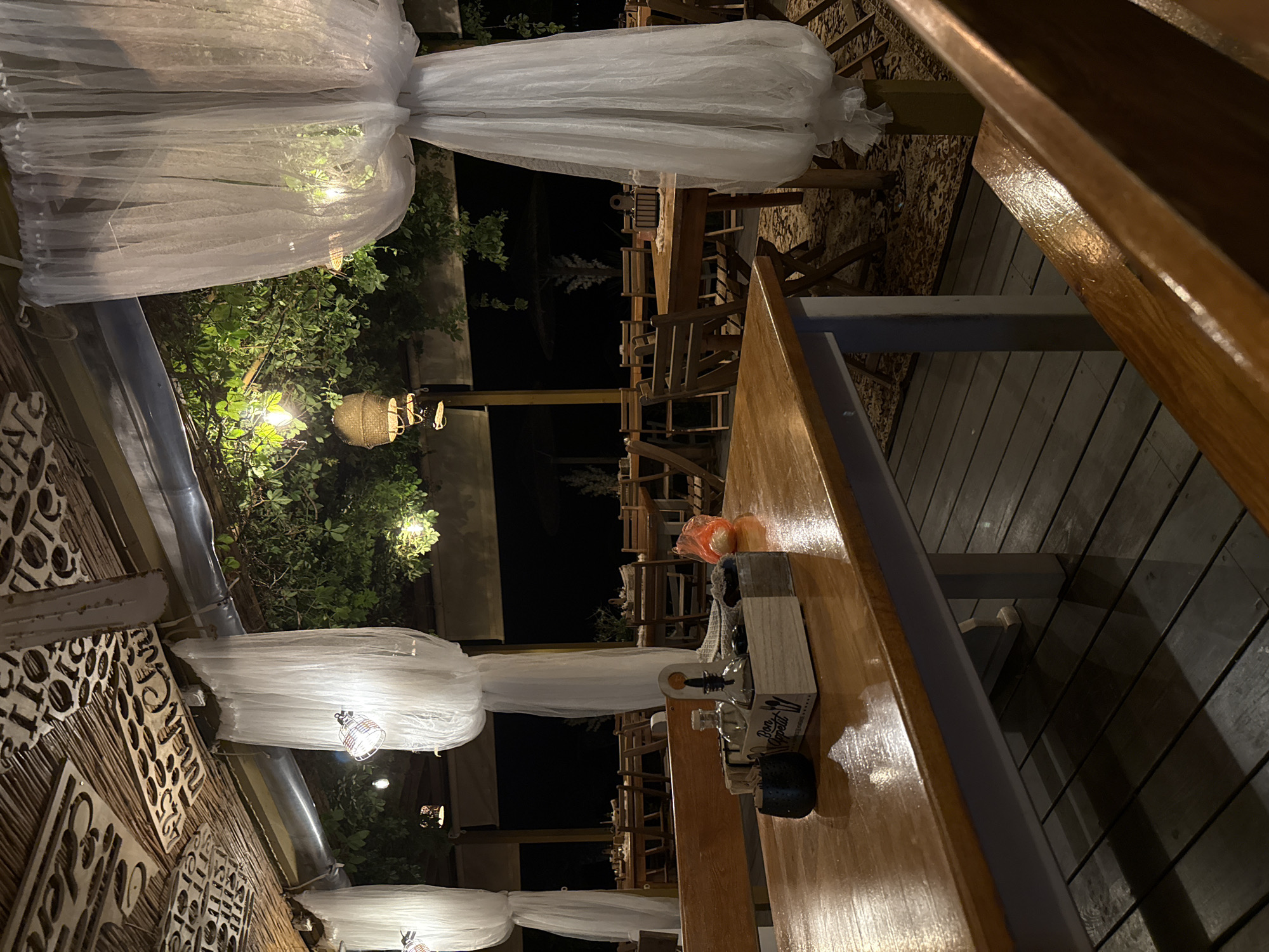 The image shows an outdoor dining area at night, likely a restaurant or cafe in Tsarevo, Bulgaria. The setting is covered by a roof structure decorated with what appears to be patterned wood panels and some text. White sheer curtains are draped between pillars, creating a soft, ethereal atmosphere. String lights and lanterns are visible, illuminating the space and casting a warm glow on the wooden tables and chairs.
In the foreground, a wooden table is set with a condiments caddy containing bottles of oil and vinegar, a napkin holder, and a small dark object, possibly a salt or pepper shaker. The table surface is polished and reflects the ambient light. Beyond the table, rows of similar wooden tables and chairs are arranged, some with place settings. A patterned rug is visible on the floor under some of the tables, and a wooden deck forms the base of the dining area. Green foliage hangs from above, adding a natural element to the scene. The background is dark, indicating it is nighttime. There are no people visible in the image.