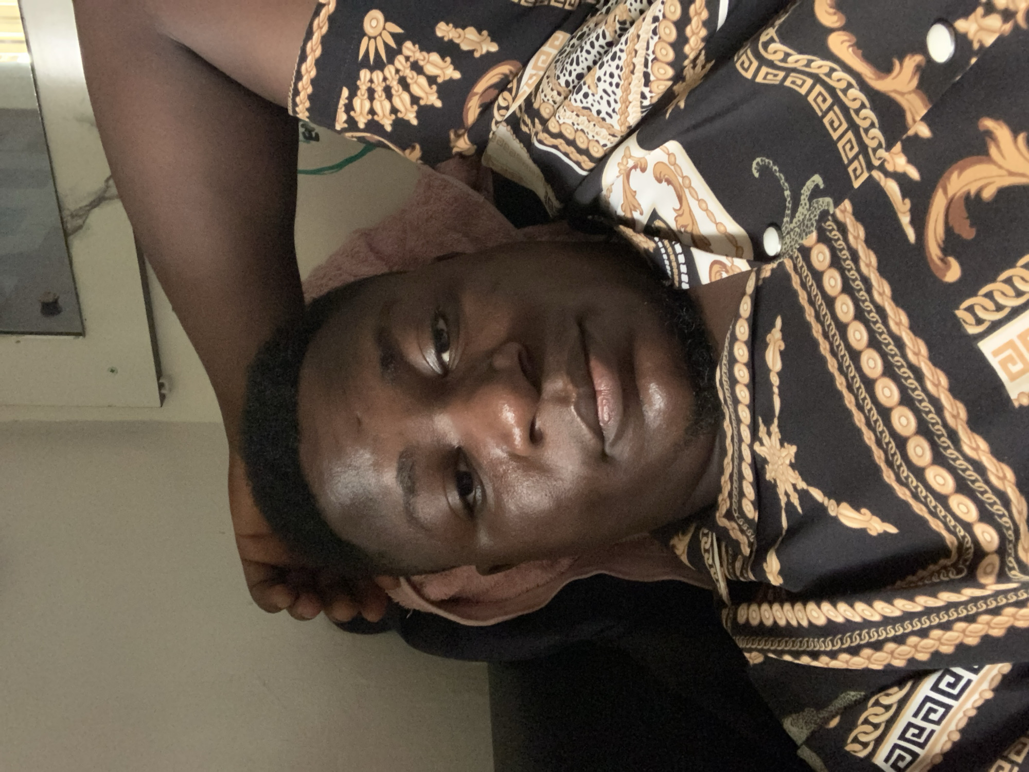The image displays a young Black man lying down in an indoor setting, looking directly at the camera. The photograph is oriented with the man's head on the right and his feet on the left, rotated 90 degrees counter-clockwise.
The man has short, dark hair and a short beard. His skin appears smooth with a natural sheen, possibly indicating warmth or hydration. He maintains a neutral and composed expression.
He is wearing a striking black shirt or robe patterned with elaborate gold and white designs. These patterns include ornate chains, scrolls, and geometric shapes, giving the garment a luxurious and detailed appearance. A light pink or beige towel is visible supporting his head and neck, suggesting he is resting on it.
To the left side of the frame, his bare legs and feet are visible, indicating he is barefoot. The background features light-colored walls and a dark, possibly reflective, surface to the far left. The lighting appears to be from an indoor source, casting a soft illumination on the scene. There is no visible text in the image. The location is contextualized as Akwanga, Nigeria.
