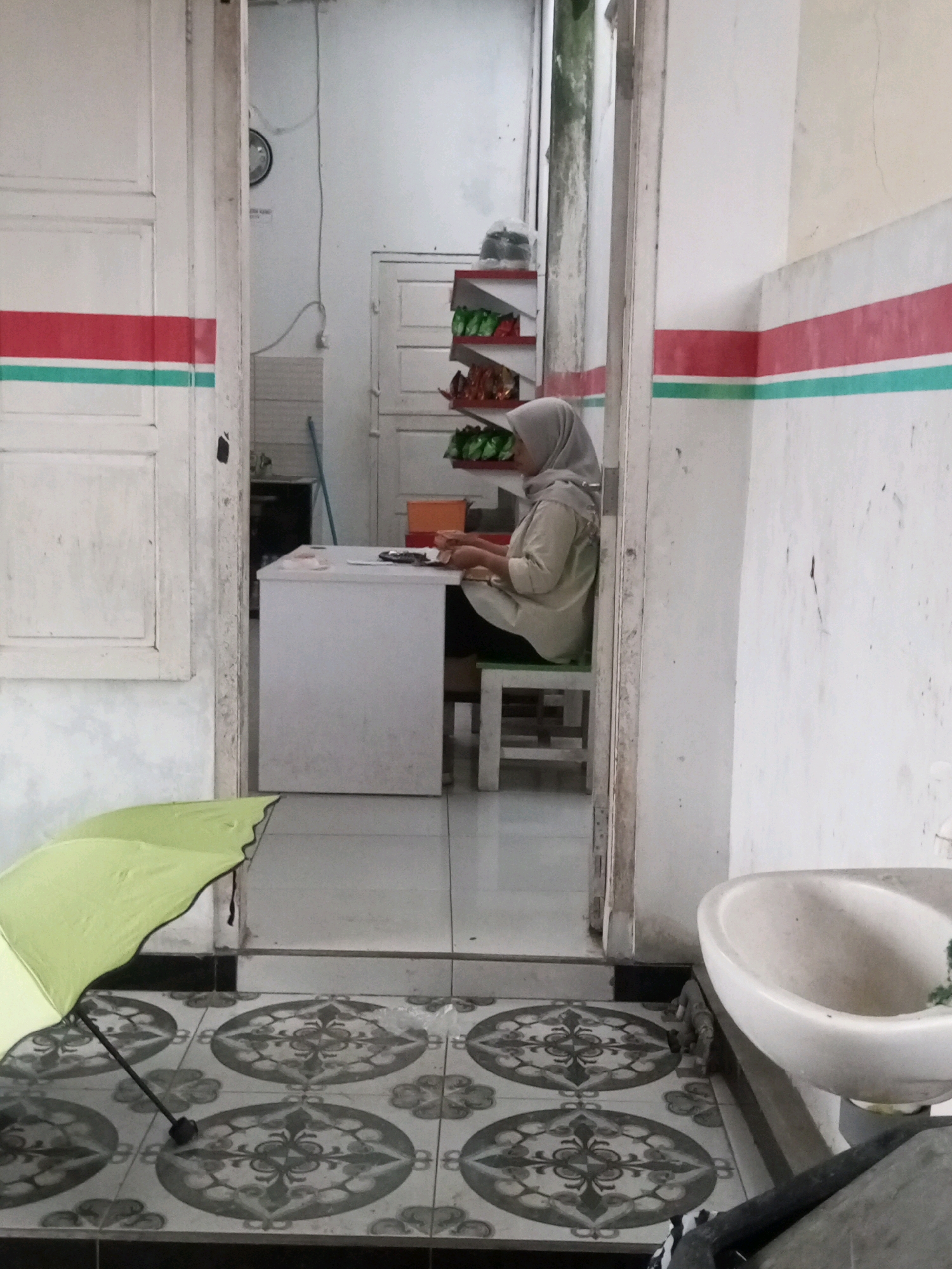 A woman wearing a hijab and a light-colored shirt sits at a white desk in a room that appears to be an office or reception area in Kalibeber, Indonesia. She is looking down at papers on the desk and appears to be working. To her left, an open doorway reveals a glimpse of another room with shelves stocked with items. In the foreground, a bright green umbrella lies on the floor. The room features tiled flooring with a decorative pattern and a sink visible on the right side of the image. The walls are painted white with horizontal red and green stripes.
