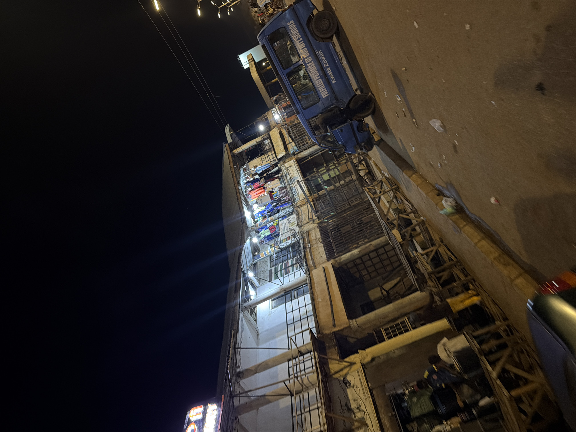 The image depicts a nighttime street scene in Bukuru, Nigeria. A blue van with "FULFILLED PROMISE OF GOD INT'L SCHOOLS" and "Kwata Zawani" written on its side is parked on the right side of the road. In the background, a two-story building with an open market on the first floor is visible. Clothes are displayed for sale on the first floor. The street is unpaved and littered with trash. Several people are standing near the market stalls. The sky is dark, and streetlights are visible in the distance. The overall impression is of a busy, lively marketplace at night.