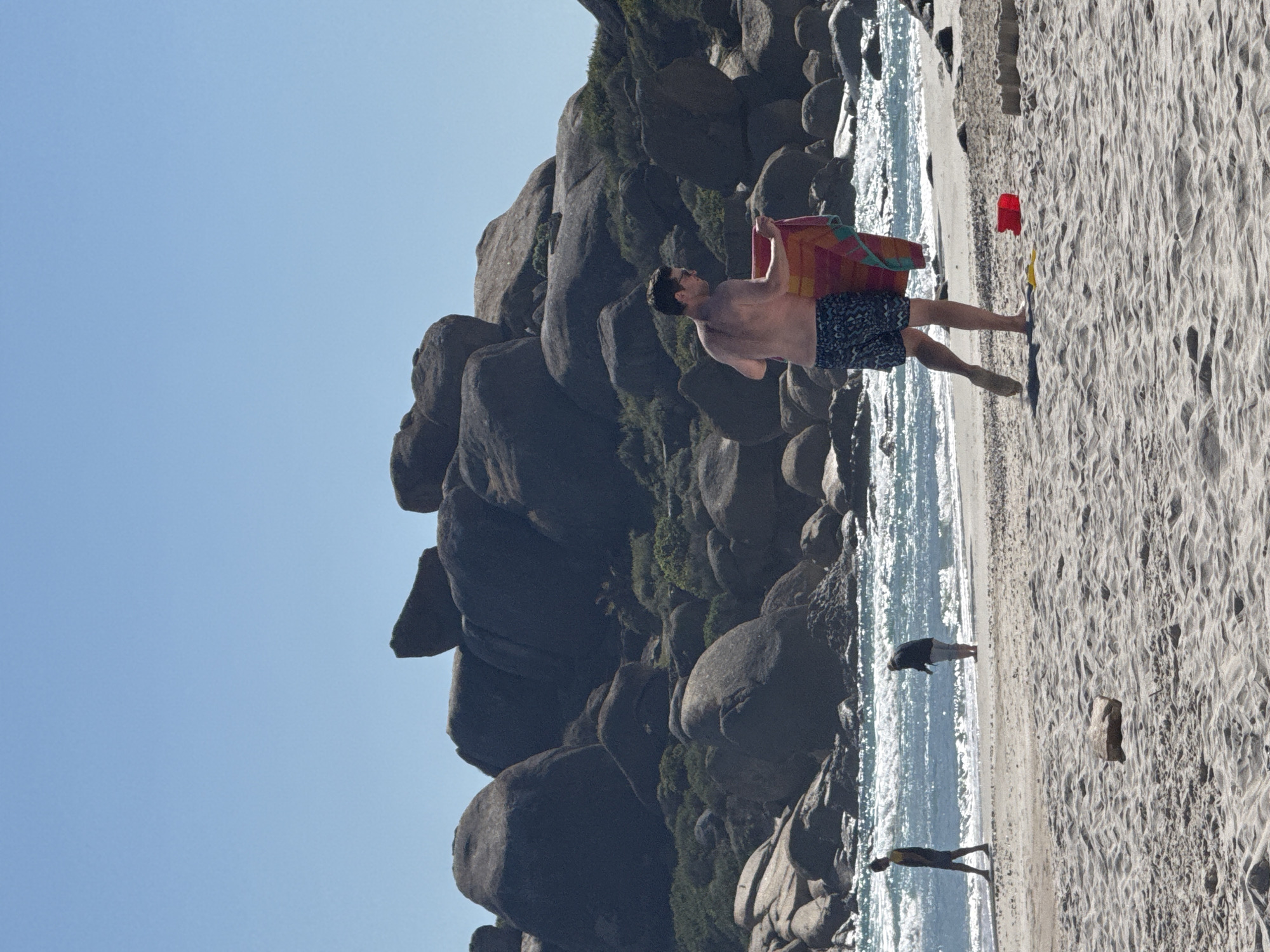 The image displays a vibrant beach scene on a sunny day, likely in Hout Bay, South Africa. The primary subject is a man in the mid-ground, walking towards the left of the frame. He is shirtless and wears dark patterned swim trunks, carrying a brightly colored, striped towel or small bodyboard (red, orange, yellow) over his shoulder.
The setting features a rugged coastline on the left side, dominated by massive, smooth, dark grey boulders and rock formations, with sparse green vegetation clinging to them. To the right of the rocks, the ocean meets the shore, sparkling under the bright sunlight. The wide expanse of light-colored sand shows clear ripple patterns, suggesting wind or wave action. Further along the beach, two other human figures are visible; one stands in the shallow water, and another is closer to the sand. Abandoned on the sand are a bright red bucket and possibly a yellow shovel, indicating leisure activities. A piece of driftwood also lies on the sandy surface. The sky is clear and a brilliant blue, characteristic of a bright daytime scene. There is no visible text in the image.