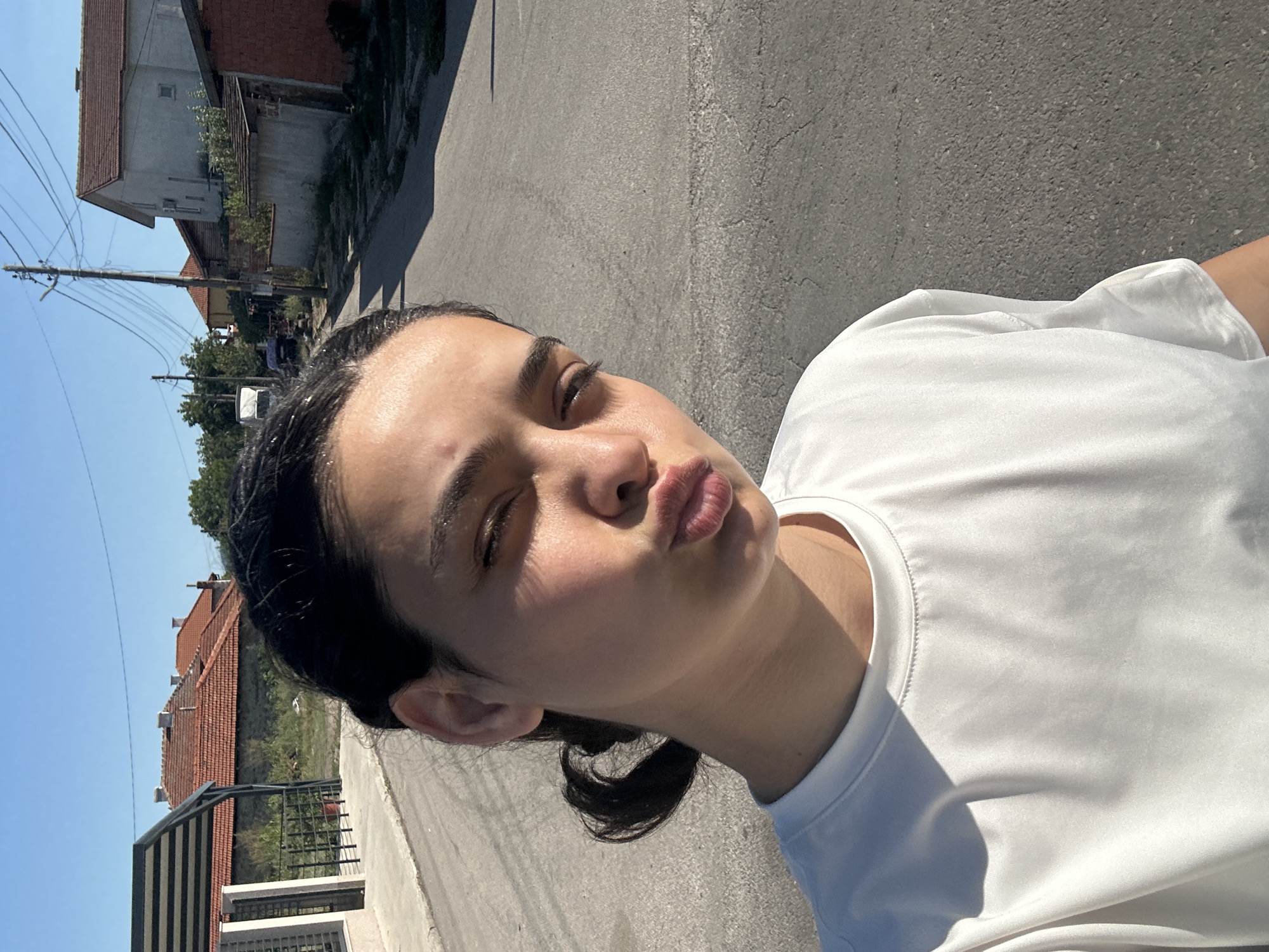 A young woman is taking a selfie outdoors on a street in Malevo, Bulgaria. She has dark hair pulled back in a low ponytail and is wearing a white t-shirt. Her face is partially obscured by the angle of the shot, but she is puckering her lips as if for a kiss. The background shows a residential street with houses, rooftops, utility poles, and trees under a clear blue sky. The sun is casting strong shadows, indicating it is likely late afternoon.