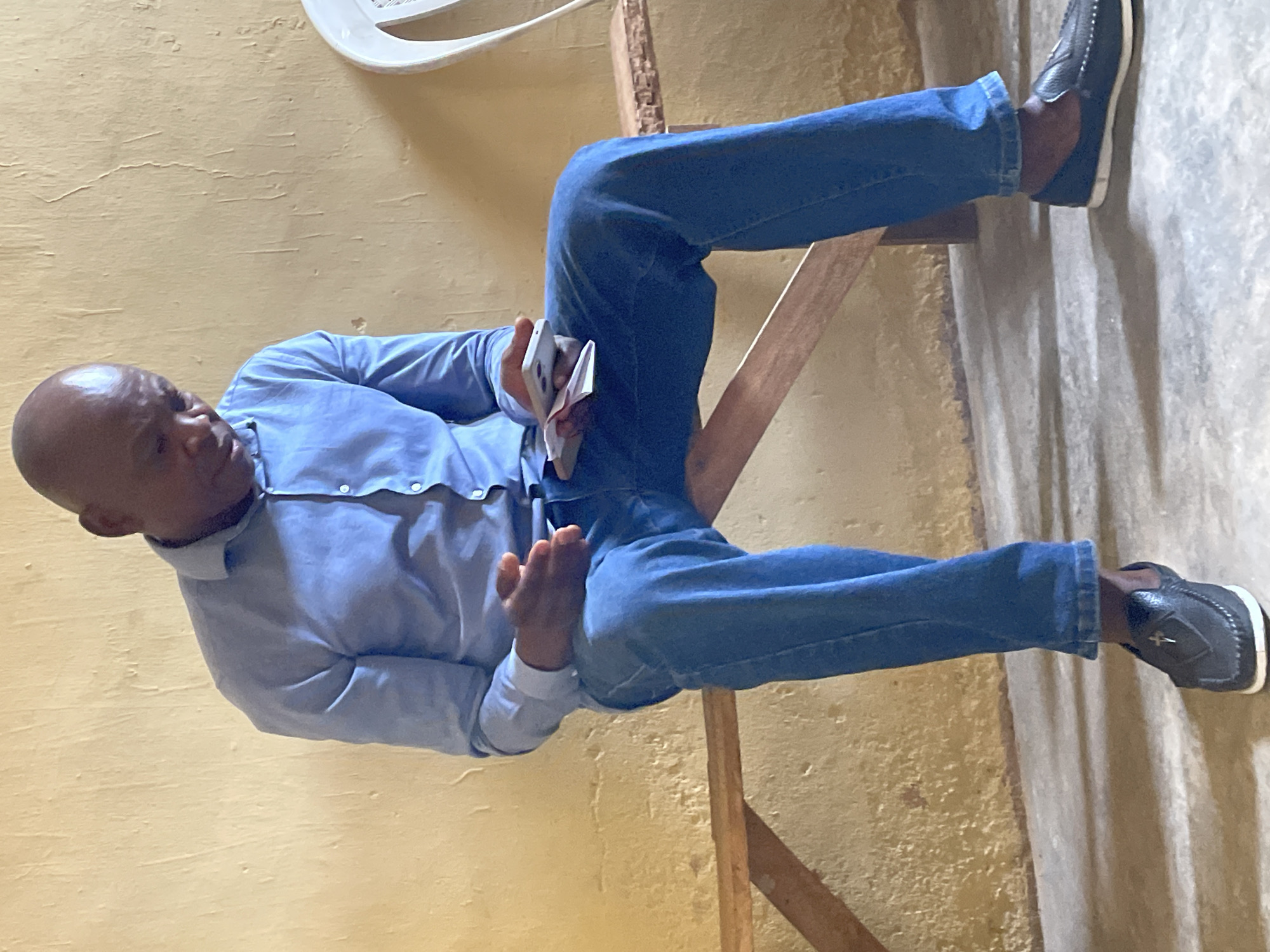 A man of dark complexion is seated indoors, facing slightly to his left with a pensive or neutral expression, looking downwards. He is bald or has very short hair. He is wearing a light blue long-sleeved button-up shirt and blue jeans. His footwear consists of dark loafers with white soles.
In his left hand, the man holds a white smartphone, partially alongside what appears to be a small stack of papers or a booklet. His right hand rests casually on his right thigh, fingers slightly curled. He is seated on a simple wooden structure, possibly a chair or stool.
The background features a textured, yellowish-beige wall that shows signs of wear or an uneven surface. The floor is a light grey, appearing to be concrete. A white, possibly plastic, object is partially visible in the upper part of the image, possibly an architectural feature or another piece of furniture. The setting appears to be a modest, casual indoor environment, consistent with the provided location context of Jos, Nigeria. There is no visible text in the image.