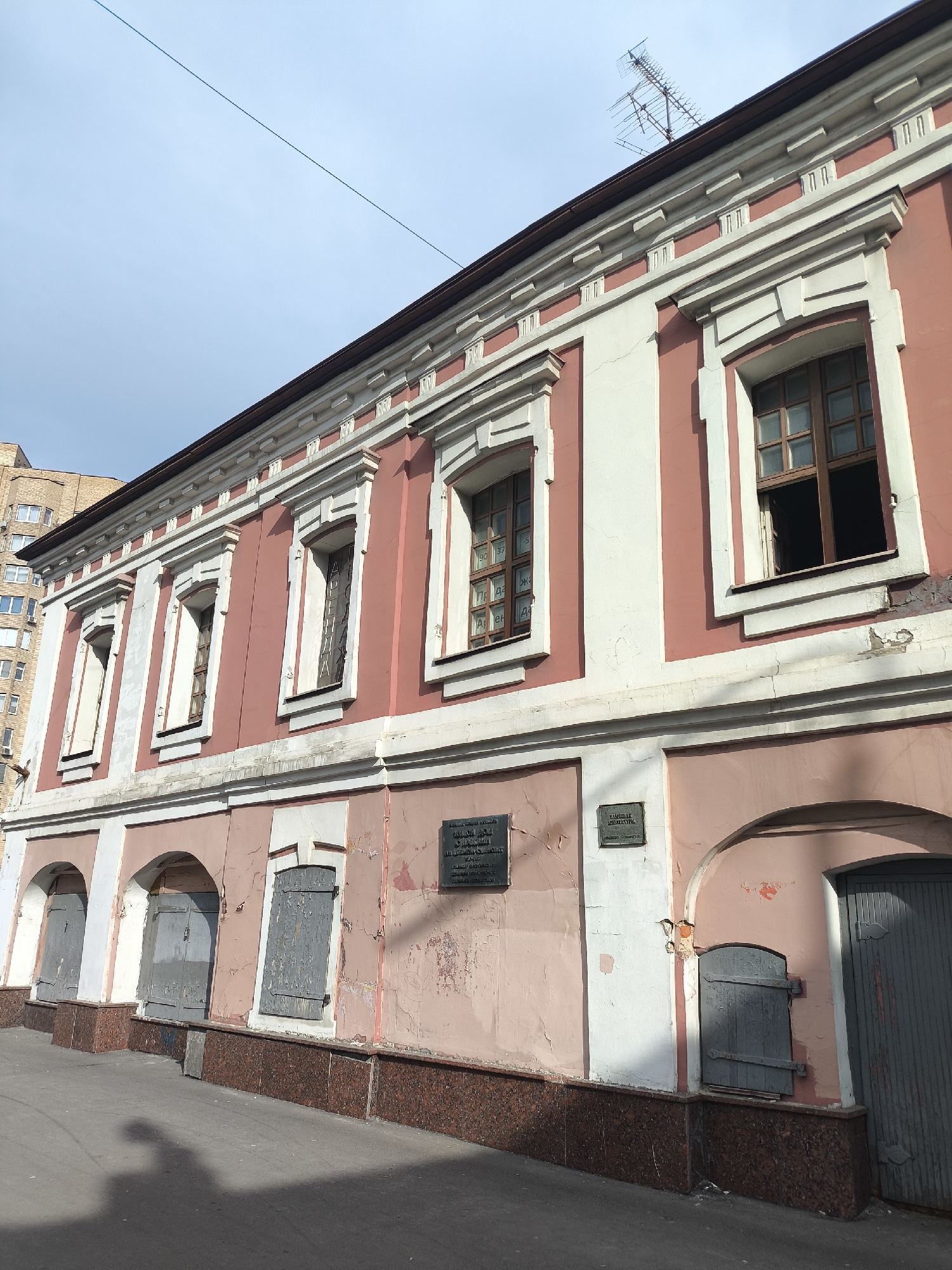 The image shows a two-story building with a pink and white facade. The upper story has several windows, some of which are open. An antenna is visible on the roof. The lower story has arched entrances, most of which are closed with wooden doors or shutters. There are two dark plaques mounted on the wall between the arched entrances. The building has a dark brown base. The image appears to be taken in Moscow, Russia, based on the architectural style and a sign in Russian language. The sky is pale blue. A shadow of a person is seen on the pavement.