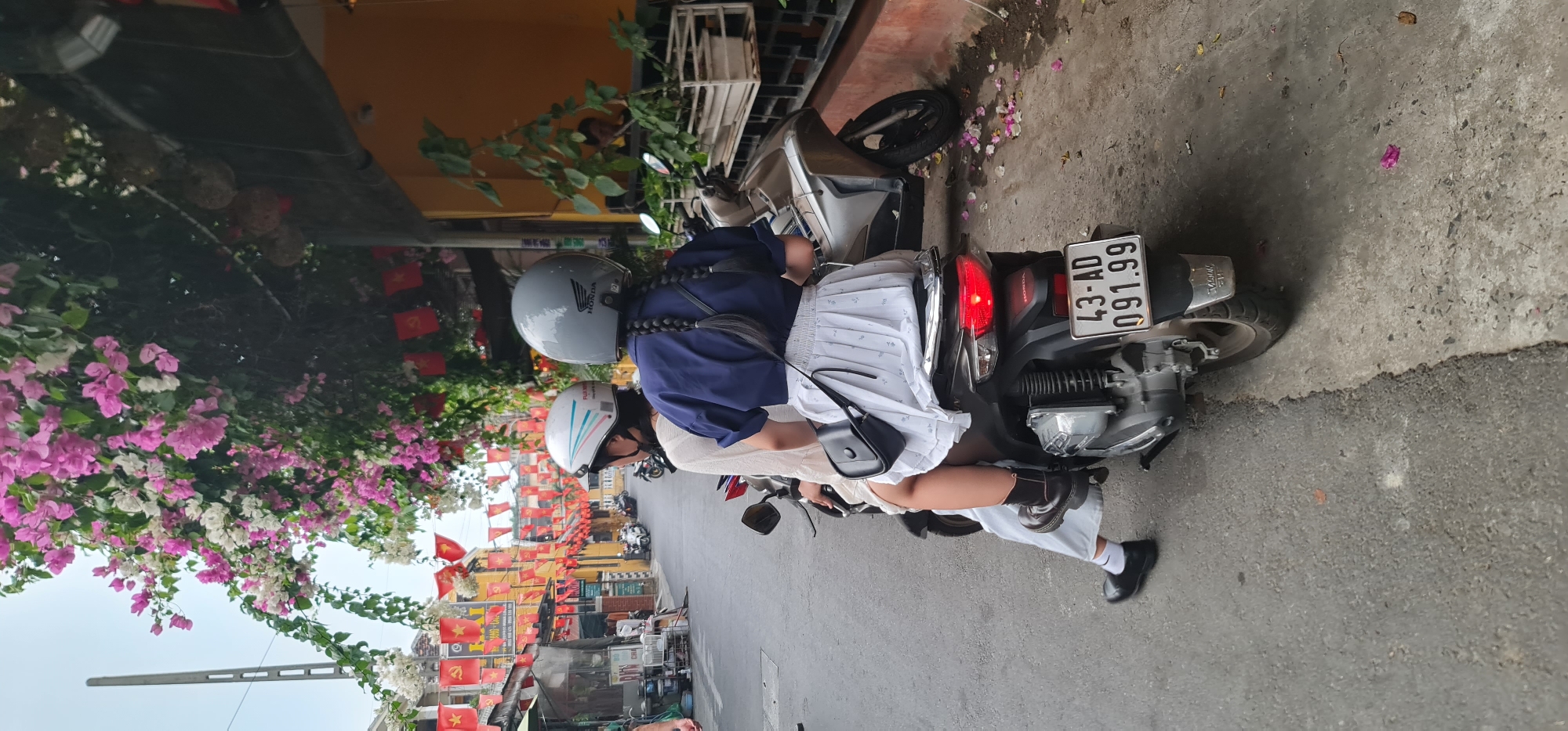 The image shows two people on a motorcycle in a street decorated with Vietnamese flags, likely in Hội An Ward, Vietnam. The motorcycle has the license plate "43-AD 091.99". The driver wears a grey helmet and a dark blue shirt with braided hair, while the passenger wears a white and blue helmet, a white dress, and black shoes with white socks. Pink and white flowers hang above the street. The background shows buildings and other motorcycles.