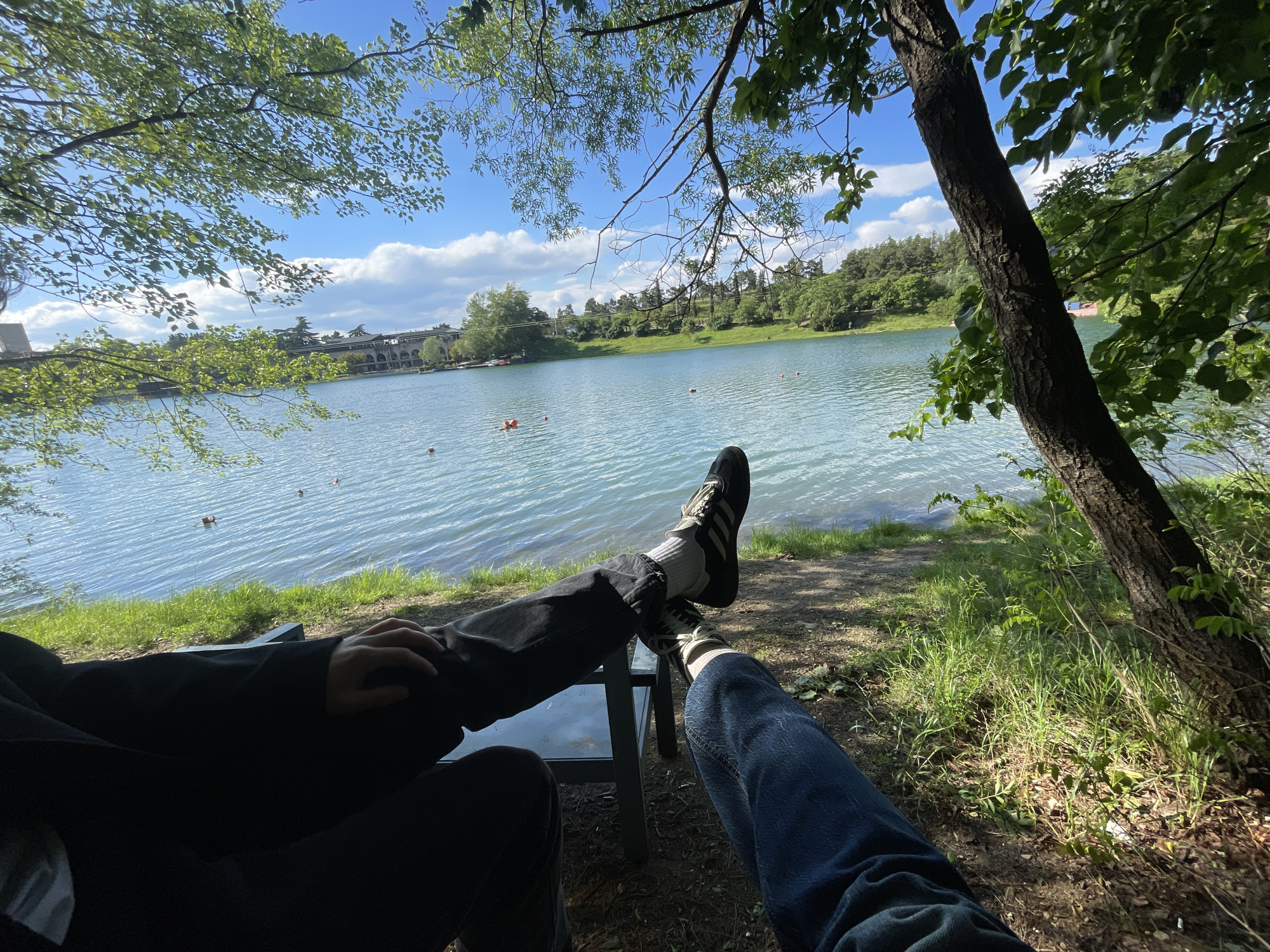 A first-person perspective shot captures a scene by a lake on a sunny day in Tbilisi, Georgia. Two individuals, likely seated, have their legs extended, feet resting on a small table. One person wears black pants and a black jacket, with black and white sneakers visible. The other wears blue jeans and white socks with black stripes, also with black and white sneakers. The legs are positioned in the foreground, with the lake filling the middle ground. Across the lake, a multi-story building with arched windows can be seen amidst green trees and vegetation. The sky is partly cloudy. Grass and a tree trunk occupy the right side of the frame.