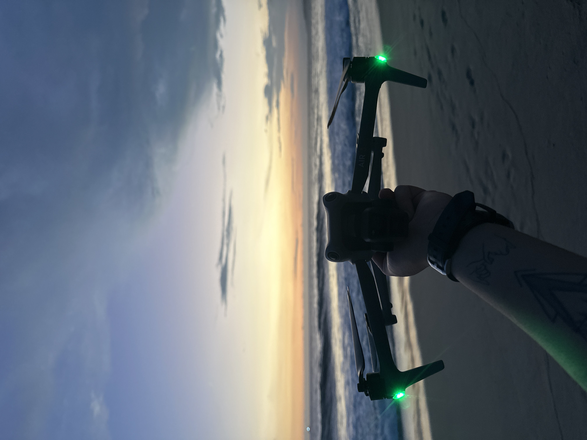 A person holds a drone with green lights on the beach at Brisas de Zicatela, Mexico. The beach and the ocean are behind the drone. The sky is visible. The person's arm has a tattoo on it. The person is also wearing a watch.
