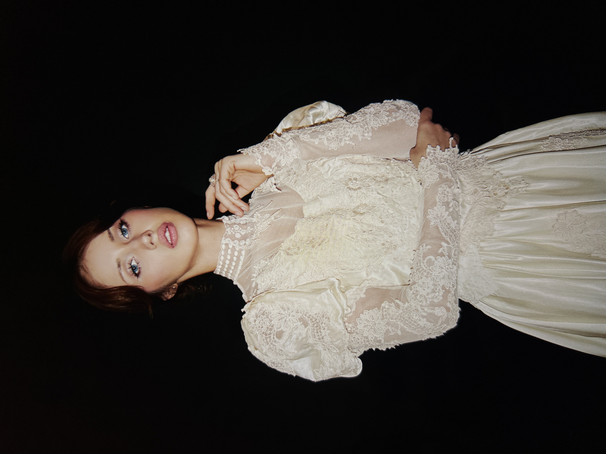 A young woman with auburn hair and blue eyes, wearing an intricate ivory lace and satin Victorian-style gown, poses against a black backdrop. Her left hand is raised, touching her chin, adorned with a ring. The dress features voluminous puffed sleeves, a high lace collar, and a full skirt with delicate floral lace appliques. The lighting is soft, highlighting the textures of the dress and the woman's features. The scene appears to be a studio portrait, with no discernible background details of a city or environment.