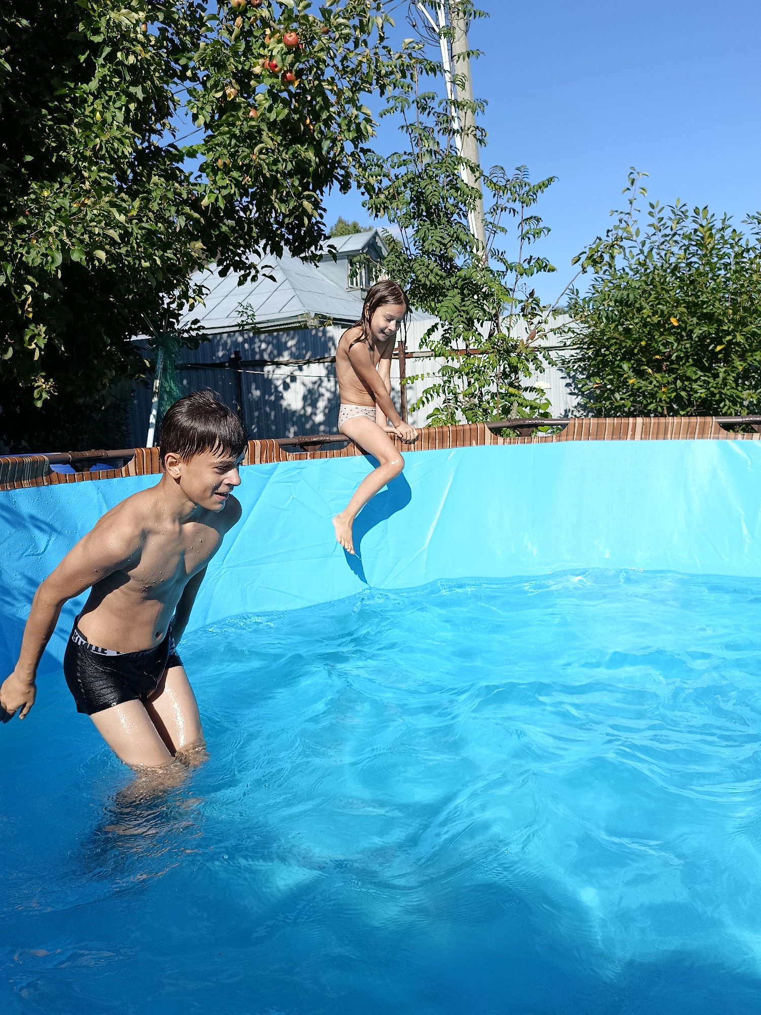 In a backyard swimming pool on a sunny day, a boy and a girl are enjoying the water. The boy, positioned in the foreground, is partially submerged in the bright blue water, looking to his right with a wide smile. He is wearing black swim trunks with white lettering. The girl, in the background, is sitting on the edge of the pool, her legs dangling over the water. She is wearing a light-colored bikini and appears to be mid-action, possibly preparing to jump in or swing her legs.
The pool itself is a large, above-ground inflatable pool with a wooden-toned border. The water is a vibrant blue and has ripples, indicating movement. Surrounding the pool, lush green trees provide shade, with some bearing red apples, suggesting late summer or early autumn. In the background, a grey-roofed building and a white fence are visible, hinting at a residential setting. The sky above is a clear, bright blue, reinforcing the impression of a warm, sunny day. The overall scene conveys a sense of playful fun and summer enjoyment. The visible text on the boy's swim trunks reads "HUMMEL".