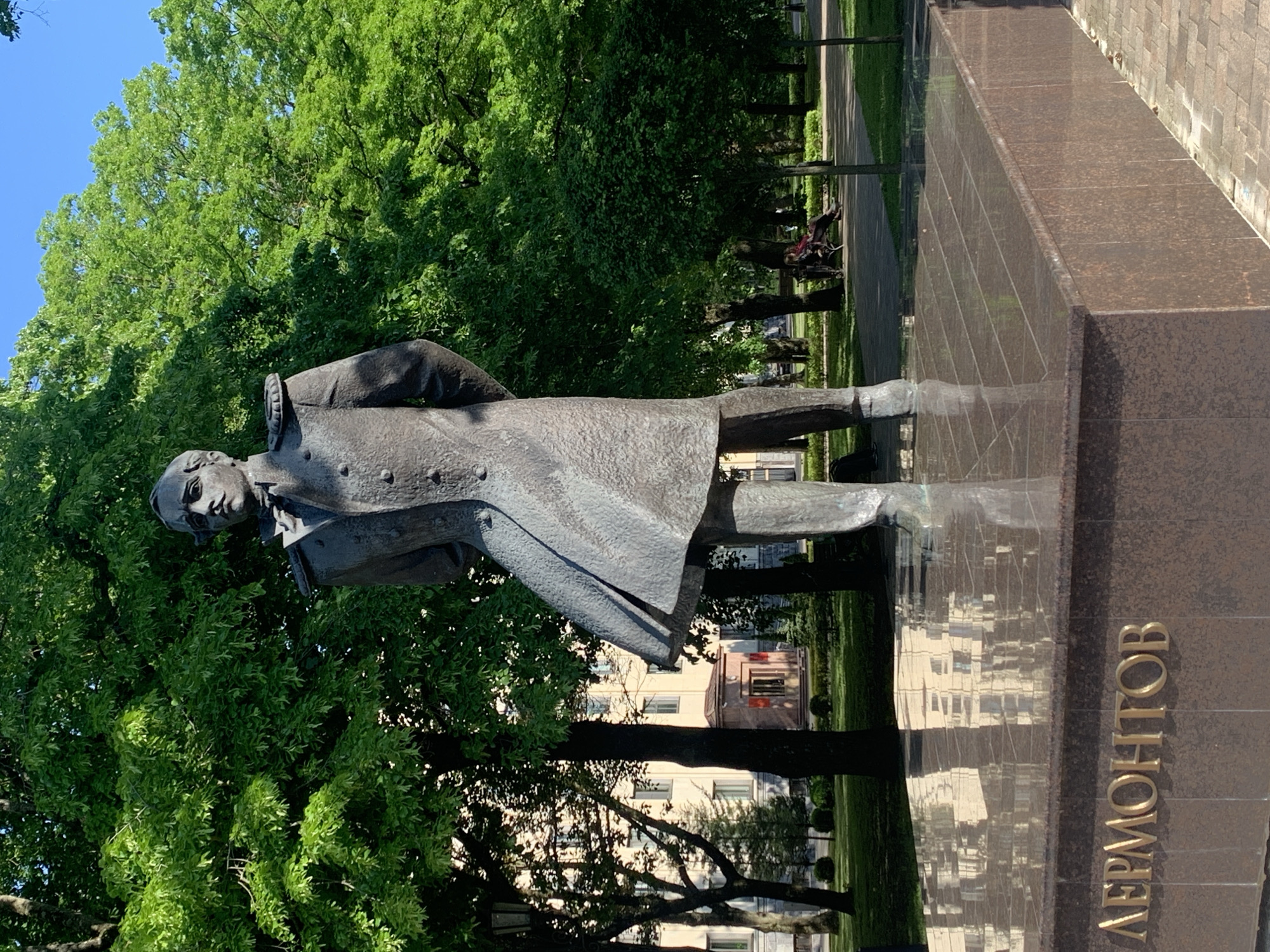 A bronze statue of Mikhail Lermontov, a prominent Russian poet and writer, stands centrally in a vibrant green park or public square under a clear blue sky. The statue depicts Lermontov in a military uniform, featuring a long, double-breasted coat with epaulets, buttons, a high collar, and a bow tie. His hands are positioned behind his back, and the lower left side of his coat is sculpted as if caught by a gentle breeze. He has a serious expression and a mustache. The statue rests on a large, polished brown granite plinth, which bears the name "ЛЕРМОНТОВ" in raised gold Cyrillic lettering on its front face. The highly reflective surface of the plinth's top shows distorted reflections of the background buildings and trees.
The park environment is lush, with numerous mature trees displaying dense, bright green foliage, some casting shadows. In the left background, partially obscured by trees, is a light-colored, multi-story building with multiple windows. Further right, a smaller structure with a reddish roof and an entrance featuring a red wall is visible, also partially hidden by trees. A paved pathway extends into the midground on the right side of the image, where at least one person, dressed in dark clothing, is seated on a park bench under the shade of trees. The sky above the trees on the right is clear and bright blue, indicating a sunny day. The precise location is in the city of Unknown, Unknown.