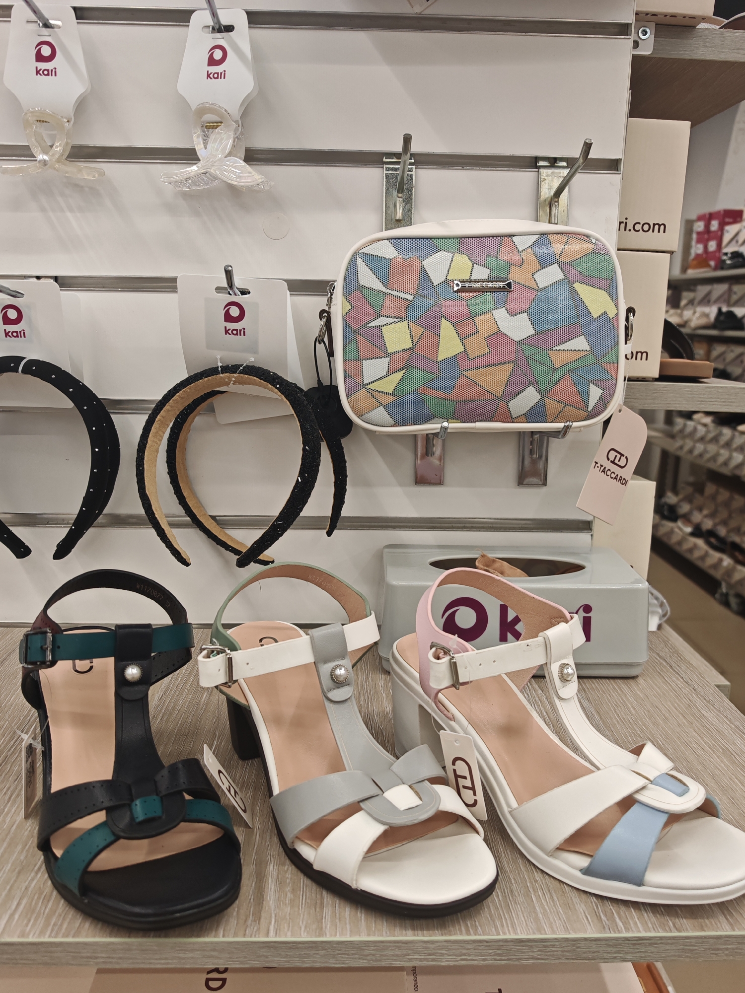 This media file is a close-up, eye-level shot of a retail display featuring women's accessories and shoes. The scene is set within a store, likely a shoe or fashion boutique, indicated by the organized display shelves and branded packaging.
The primary subjects are various fashion items. On the upper shelves, there are clear plastic hair clips with iridescent finishes, each attached to white cardboard tags bearing the "Kari" logo. Below these, two black headbands, one adorned with small rhinestones and the other with a textured beaded pattern, are displayed.
In the center of the frame, a small, rectangular handbag with a colorful geometric mosaic pattern is showcased. It hangs from a metal hook. Adjacent to the handbag, a light gray tissue box is visible, with a pink and white sandal strap extending over it.
The lower shelves display three pairs of women's sandals. The leftmost sandal is black with teal accents and a T-strap design. The middle sandal is white with light gray and white straps. The rightmost sandal is white with light blue and white accents. All shoes are presented on a light wood-grain surface.
Behind the handbag and tissue box, white cardboard boxes are stacked, one of which has "ri.com" printed on it. A pink tag with a "T-TACCARDI" logo is also visible. The lighting appears to be artificial, typical of an indoor retail environment, with no indications of time of day or weather. The overall impression is one of commercial display, focusing on the presentation of merchandise.