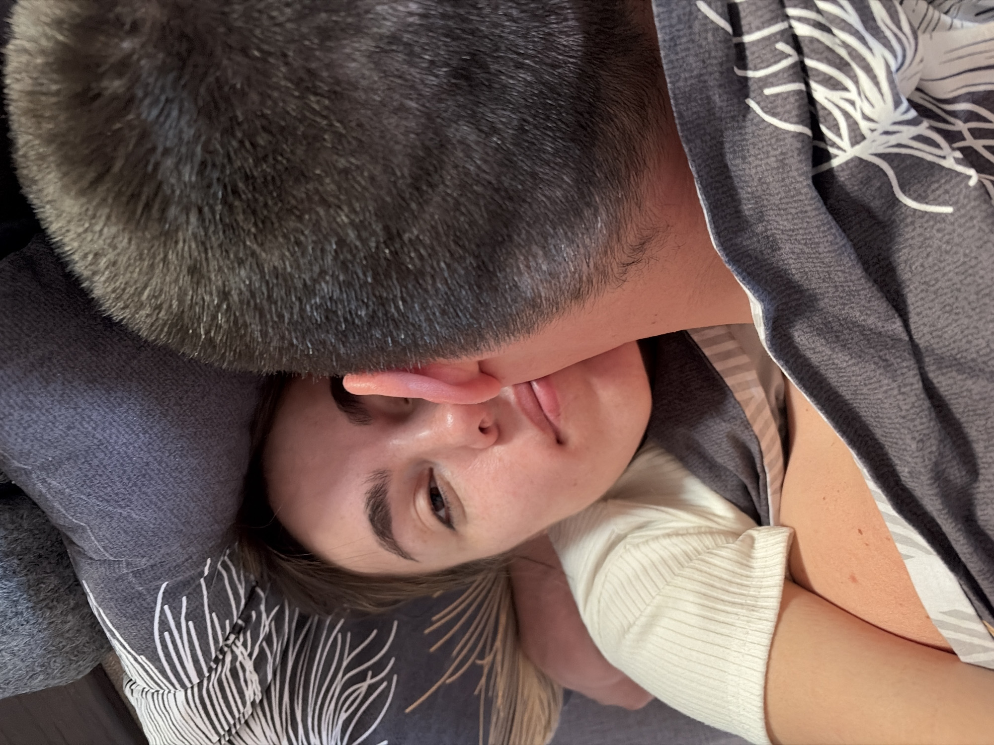 The image shows a woman lying down with her head on a patterned gray pillow and covered with a gray and white patterned duvet. A person with short, dark hair is close to her face. The woman is wearing a white top. The location is not discernible from the image, and therefore cannot be identified as Izhevsk, Russia.
