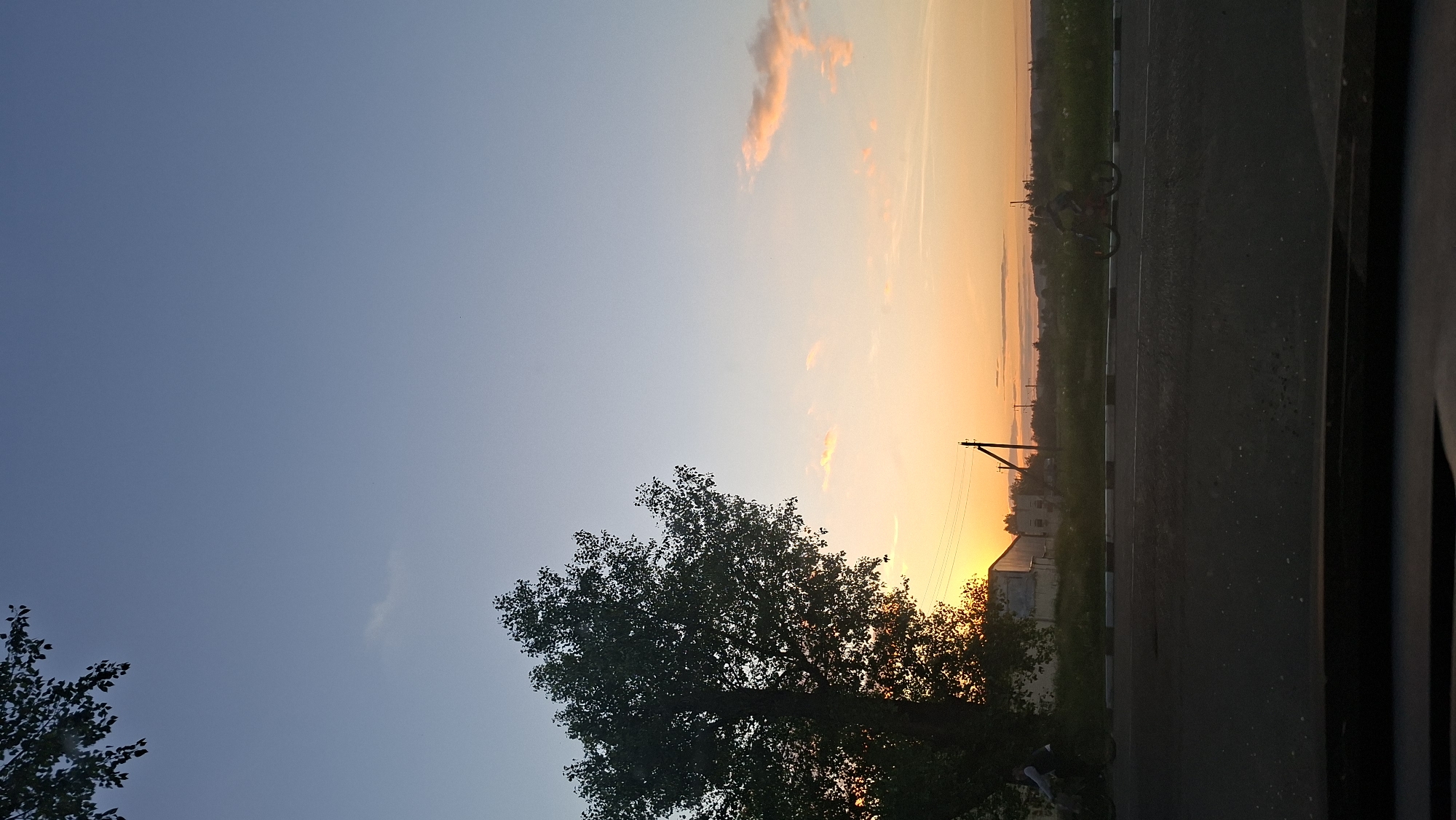 The image depicts a serene landscape at either sunrise or sunset, characterized by a dramatic sky. The sky transitions from a clear, deep blue at the top to vibrant orange and yellow hues near the horizon, indicative of a low sun. A few wispy, light-colored clouds are scattered across the orange portion of the sky, along with faint linear contrails.
In the foreground, a large, dark tree with dense foliage is silhouetted against the colorful sky, dominating the left side of the frame. To the right, a dark asphalt road runs horizontally across the lower part of the image, bordered by a white broken line and a dark curb. Beyond the road, a strip of green terrain, possibly fields or overgrown areas, stretches towards the horizon.
In the mid-ground, a wooden utility pole with visible wires stands upright, silhouetted against the bright horizon. Further in the distance, on the left side of the road, a bicycle is faintly discernible as a dark silhouette. Several indistinct buildings or structures are visible in the background, including one light-colored building that stands out against the low sun. Near the bottom left corner, beneath the large tree, a faint, dark silhouette of what appears to be a person can be seen, possibly stooping or walking.
The scene conveys a peaceful and tranquil atmosphere at dawn or dusk, with no specific activity highlighted other than the natural transition of day to night or vice-versa. There is no visible text in the image. The setting suggests a rural or semi-urban area, consistent with the provided location context of Забалоцкі сельскі Савет, Belarus.