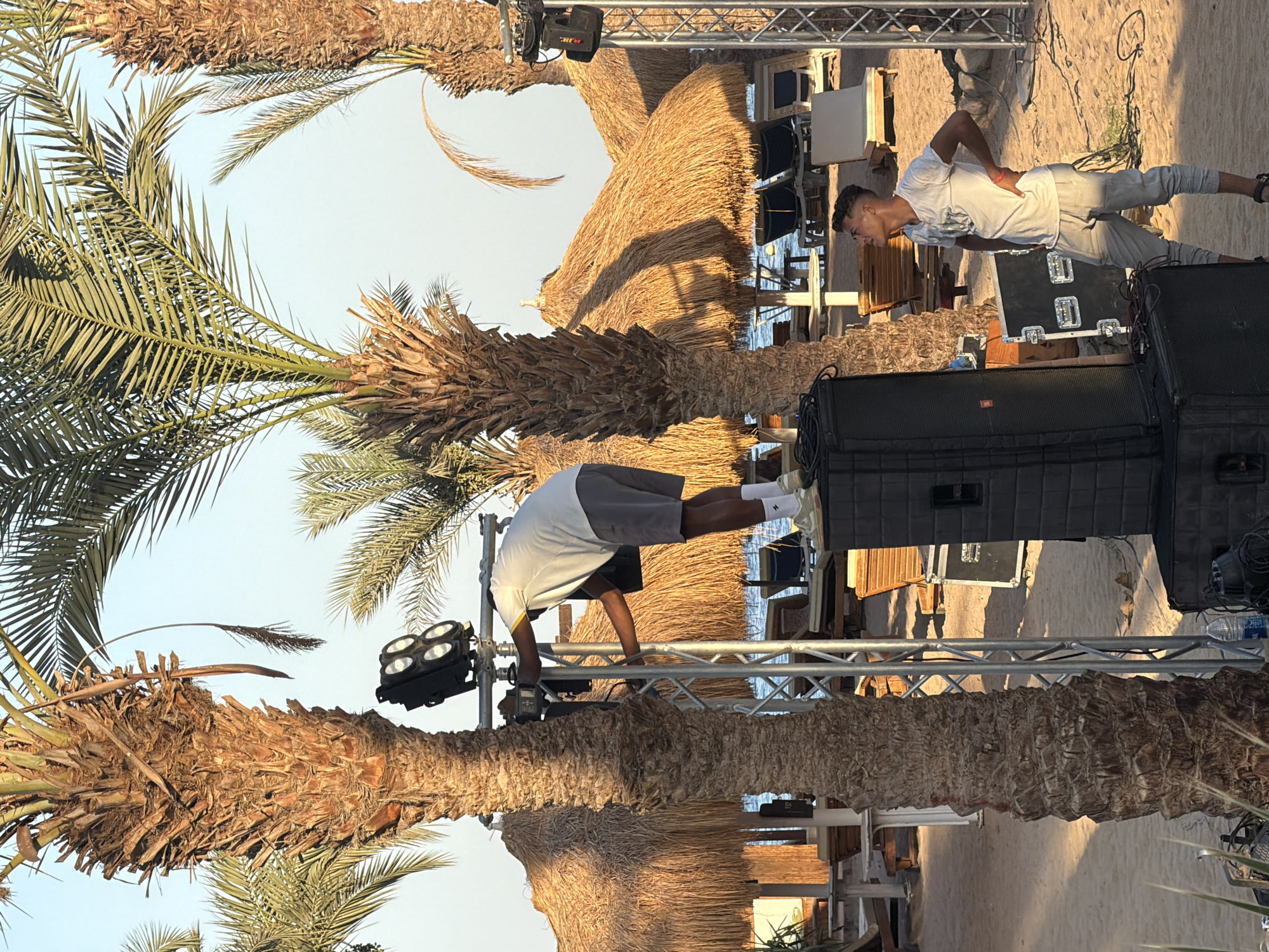 This image depicts an outdoor scene on a beach in Sharm El Sheikh, Egypt. In the foreground, two young men are engaged in setting up audio and lighting equipment. One man, wearing a white t-shirt and grey shorts, is positioned on top of a large black speaker, reaching towards a metal truss structure. He is adjusting equipment attached to the truss, which also holds four spotlights. The second man, dressed in a white t-shirt and grey pants, stands on the sandy beach near a stack of speakers, looking upwards.
The environment includes several palm trees, their trunks prominent in the frame. In the background, there are thatched umbrellas providing shade over lounge chairs on the beach, with the turquoise waters of the sea visible beyond. The lighting suggests it is daytime, likely late afternoon or early evening given the warm tones and shadows. The overall impression is of a beach venue preparing for an event.