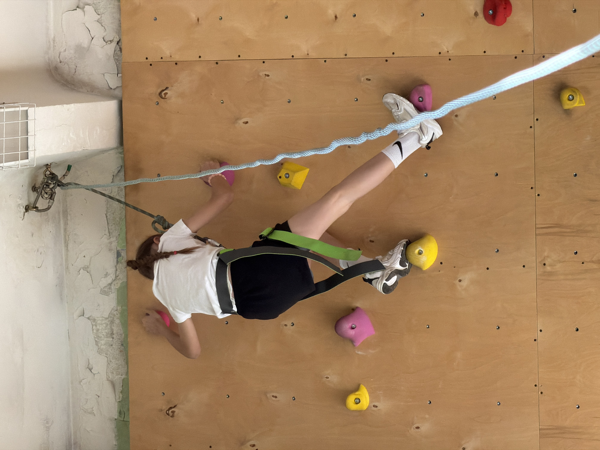 A young person is engaged in indoor rock climbing on a wooden climbing wall. The person is wearing a white t-shirt, dark shorts, climbing shoes with white socks, and a climbing harness. They are secured to a rope system that is attached to the top left corner of the wall. The climbing wall has several colorful holds attached to it. The surrounding environment includes a wall with peeling paint near the climbing wall. No specific evidence suggests a location in Izhevsk, Russia.