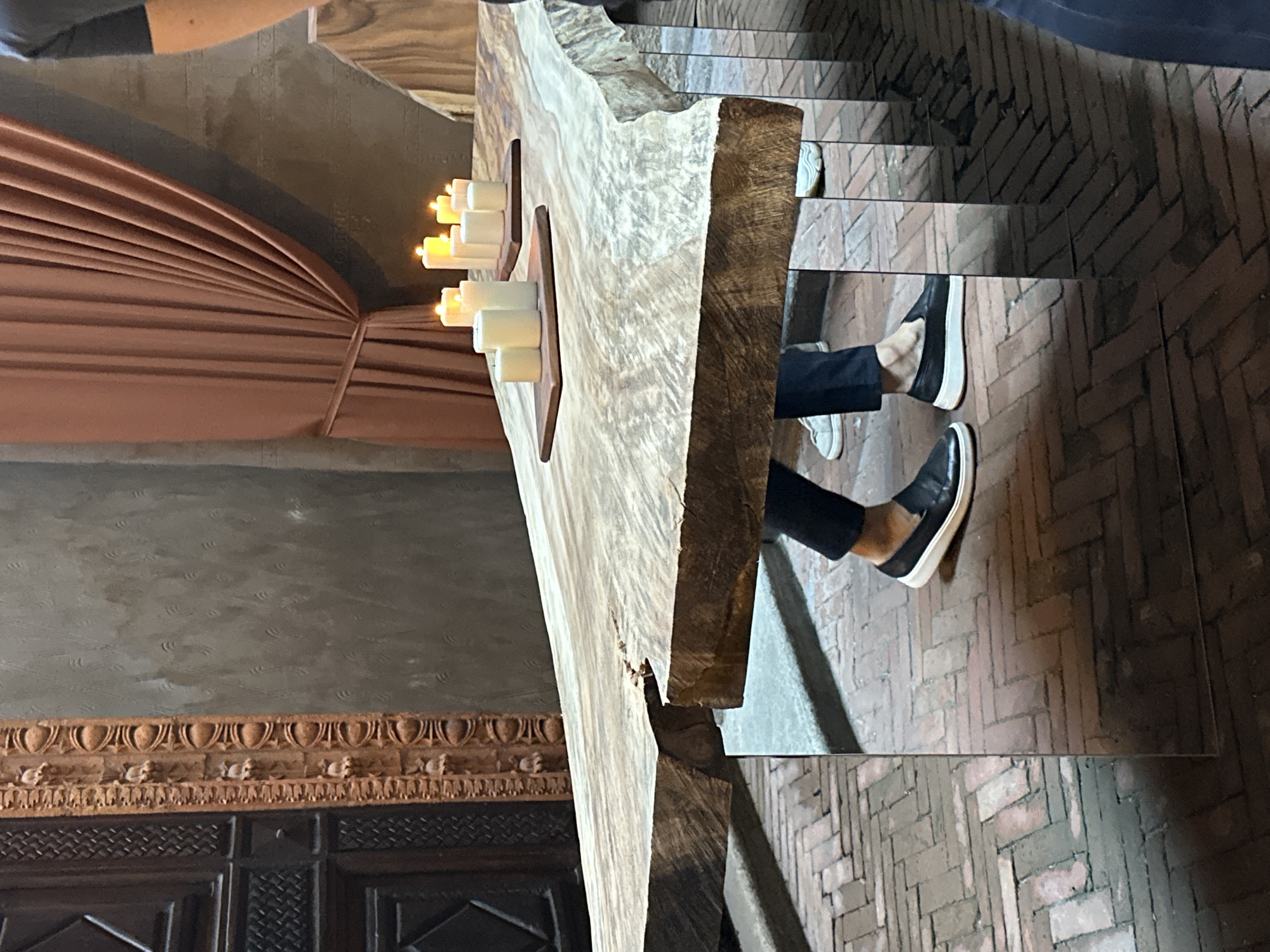 The image depicts a fragment of a scene within a dimly lit interior. In the foreground, there is a roughly hewn, large wooden slab adorned with several lit candles. To the left of the wooden slab is drapery of a pinkish-beige hue, and below it, a dark, ornately carved wooden panel. To the right of the wooden slab, a person's feet clad in black shoes are visible, seemingly situated on tiled steps. The floor is made up of bricks in a herringbone pattern. The setting suggests a somewhat medieval or rustic aesthetic. No city or country is given in the image.