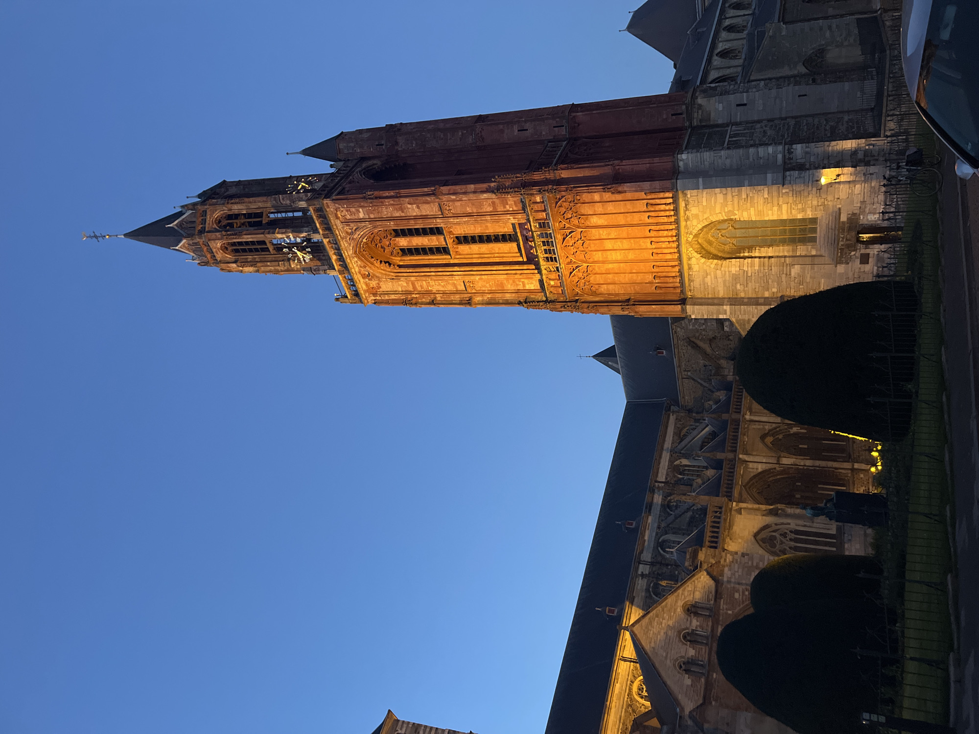Captured in the city of Maastricht, Netherlands, the image showcases the illuminated St. Servatius Basilica against a dusky blue sky. The tower of the basilica, featuring prominent architectural details, is brightly lit, contrasting against the twilight sky. Alongside the basilica, a black fence and trimmed bushes add a sense of foreground depth. A vehicle is partially visible in the upper right corner of the frame.