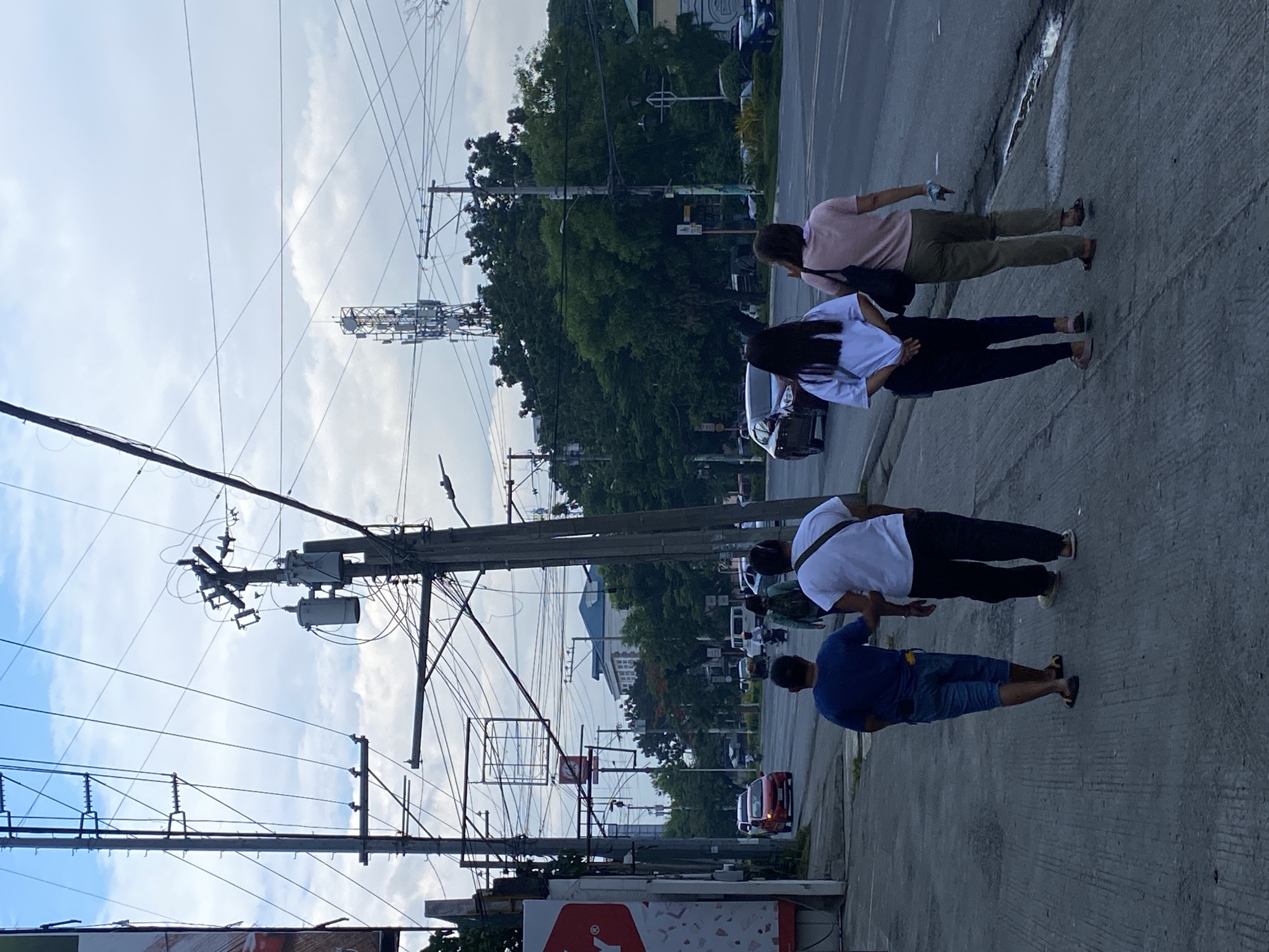 The image shows a street scene in Davao City, Philippines, with a group of people walking on a sidewalk. In the background, there are cars, a cell tower, and utility poles with many wires. The sky is partly cloudy. The group on the sidewalk consists of two men and two women. The two men are walking ahead, one wearing a blue t-shirt and denim shorts, and the other a white t-shirt and black pants. The two women are walking behind them, one wearing a white shirt and dark pants, and the other a pink shirt and khaki pants. The overall impression is of a typical urban environment with a mix of pedestrians and traffic.