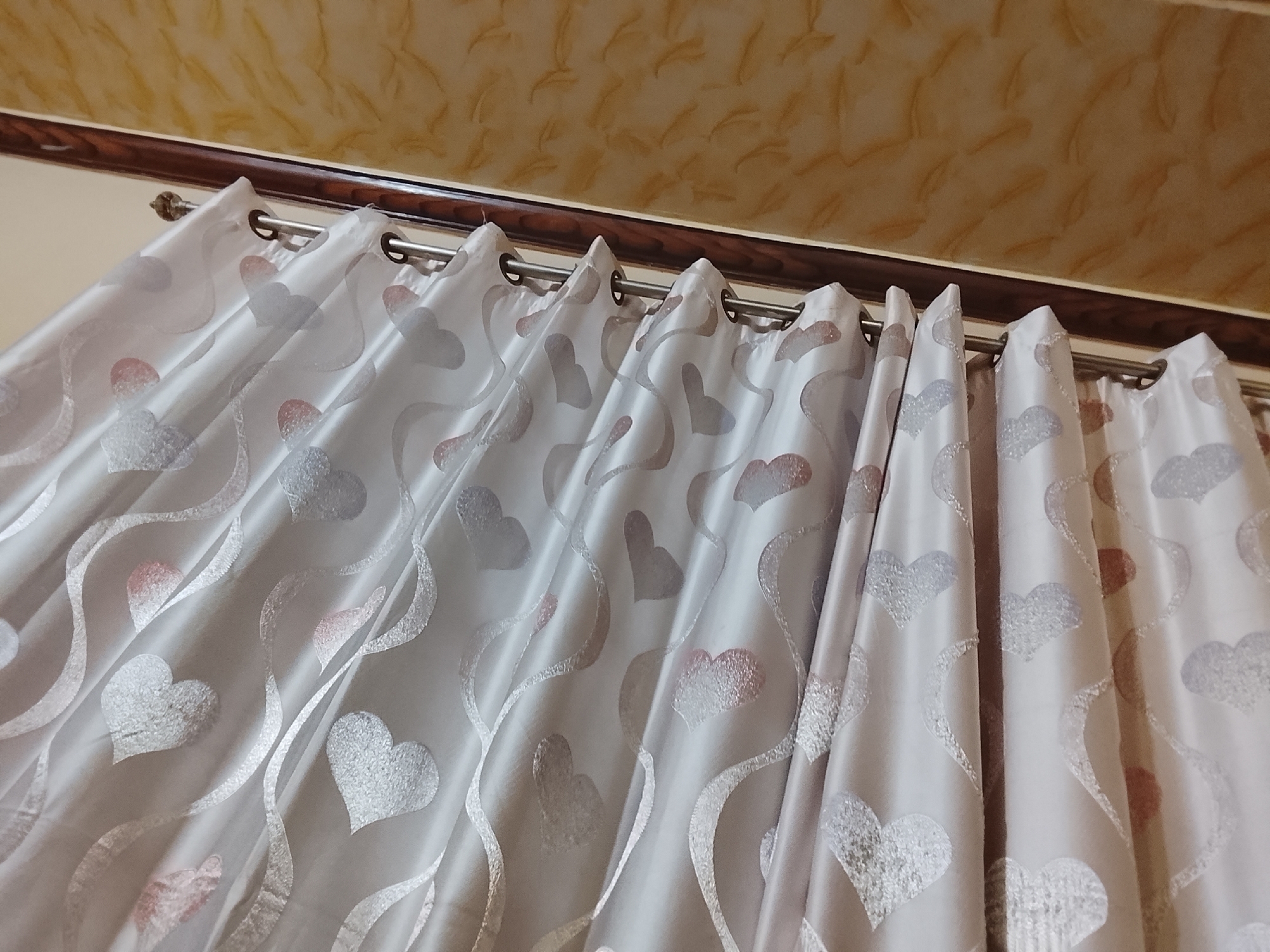 The image captures an upward view of light-colored curtains adorned with heart patterns, hung indoors. The curtains are a soft white or off-white, made of a translucent fabric, and are arranged in gentle folds. They feature a recurring motif of hearts in varying sizes, some appearing silvery and speckled, while others have a faint pinkish or reddish tint. Wavy, ribbon-like designs in a similar silvery hue are integrated throughout the heart pattern.
The curtains are suspended by silver-toned metal grommets, which thread onto a visible metallic curtain rod. Above the curtains and rod, a dark brown, polished wooden trim or valance spans the width of the frame. The uppermost section of the image reveals a light brown or tan ceiling, characterized by a distinct, repetitive textured pattern.
The scene is a static indoor shot, likely depicting a window treatment in a domestic setting. The lighting is soft and diffused, without strong shadows, suggesting ambient light rather than direct sunlight. There are no people or specific activities taking place, and no visible text is present in the image.