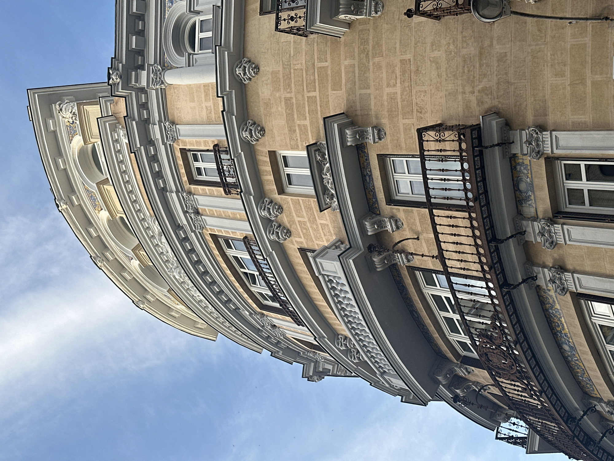 The image shows the corner of a building in Madrid, Spain, looking up towards the sky. The building has a curved facade with multiple windows and ornate architectural details, including decorative moldings, balconies, and embellished window frames. The building's exterior is painted in beige and white colors. The sky is blue with some light clouds.