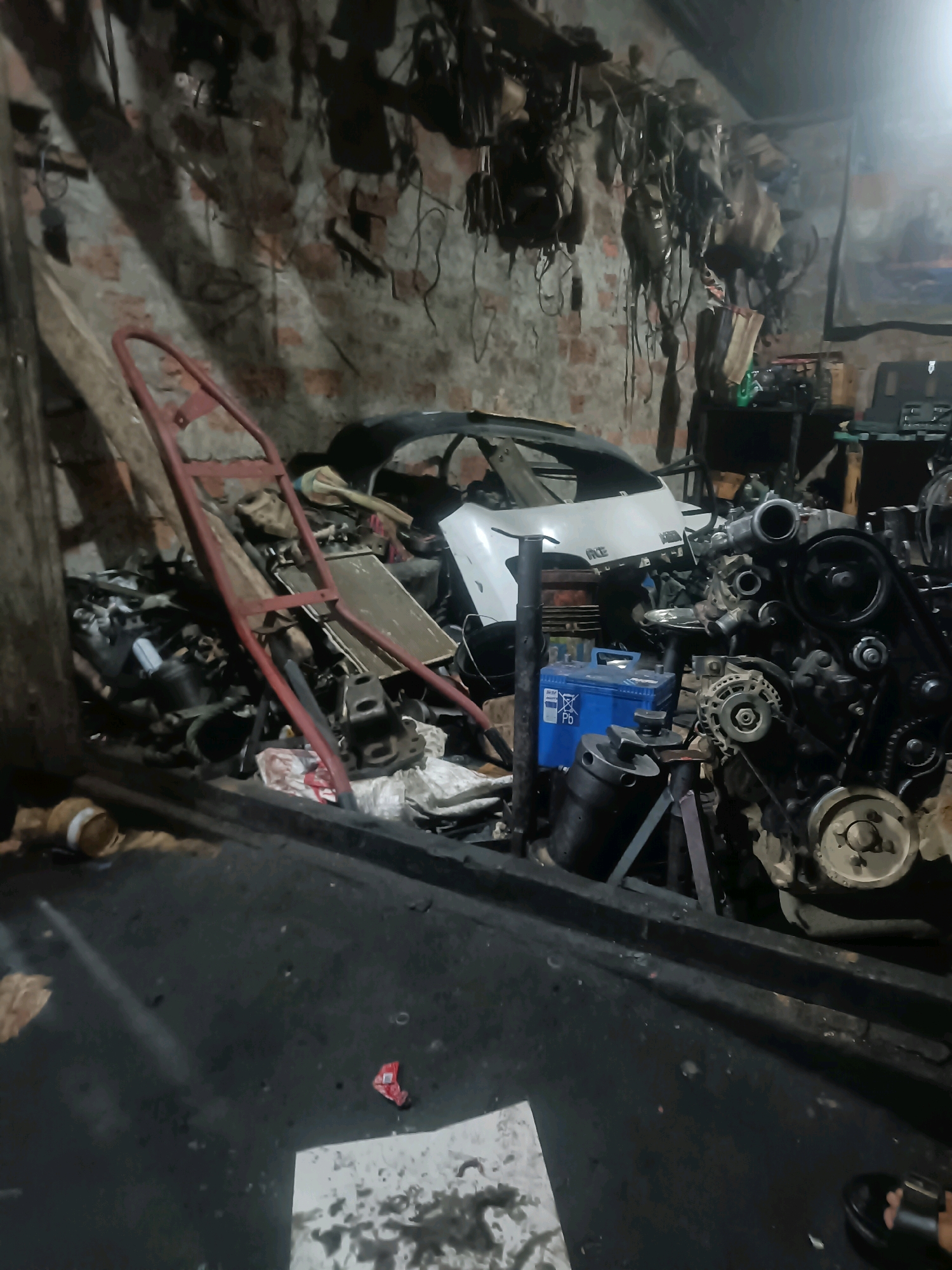 The image depicts the interior of a cluttered auto repair shop in Kuwarpar, India. The scene is dominated by various vehicle parts, including a car engine prominently displayed in the foreground with intricate mechanical details. Scattered around are other engine components, a car chassis, and a car battery. The shop's environment appears unkempt, with tools and materials strewn about. The walls are brick, and the lighting is dim, suggesting it is an indoor, possibly enclosed, workspace. There is no visible human activity or interaction captured in the frame, though a person's footwear is visible in the bottom right corner, indicating someone is present. The overall impression is one of a working garage, actively engaged in vehicle repair or maintenance, with a focus on automotive mechanics.