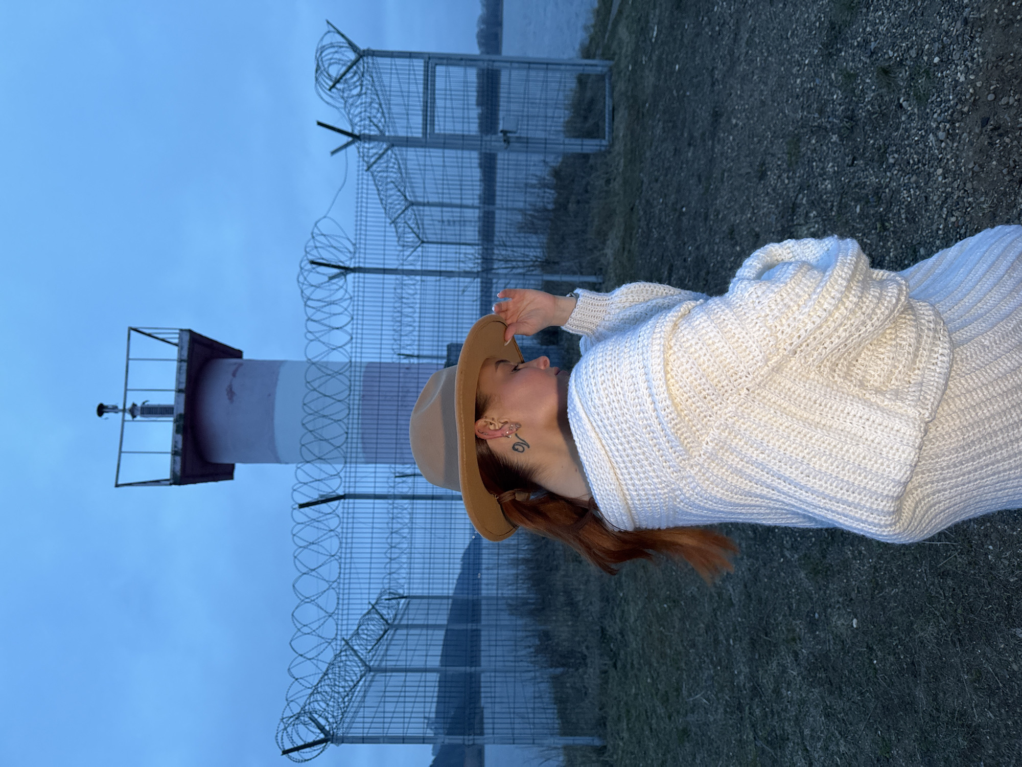 The image shows a full-length profile view of a woman standing outdoors, facing left, on what appears to be an overcast day, possibly at dusk or dawn. She is wearing a light beige fedora-style hat and a chunky knit white sweater, with reddish-brown hair visible from under her hat. Her right hand is raised, touching the brim of her hat or her temple. A small tattoo and ear piercing are visible behind her ear.
Behind the woman, a tall metal security fence topped with multiple coils of concertina barbed wire stretches horizontally across the scene. To the left of the woman and behind the fence, a lighthouse or navigation beacon with distinct white and red stripes stands. Further in the background, a wide body of water, likely a river or lake, is visible beneath a uniform blue-gray sky. The ground in the foreground consists of dark earth, small gravel, and sparse patches of grass. The lighting is soft and diffused, creating a cool, subdued atmosphere. No visible text is present in the image. The setting suggests an outdoor location near a waterway.