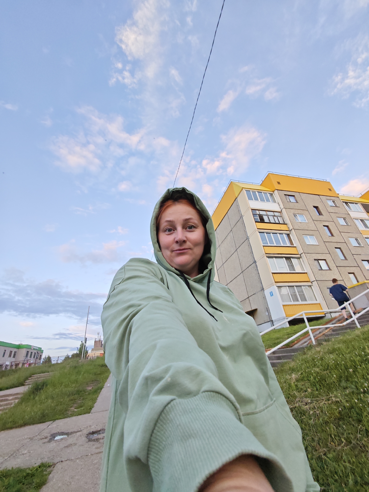 A woman with red hair, wearing a light green hoodie, takes a selfie outdoors. She is in the foreground, looking at the camera. Behind her, a multi-story apartment building with yellow and gray accents dominates the scene. A set of stairs with white railings leads up to the building, and a person in a dark shirt is visible ascending the stairs. The background consists of a cloudy sky with a hint of pink, a grassy slope, and a concrete path. In the distance, another building with a green roof is visible. The overall setting appears to be a residential area in the city of Markovsky, Russia.